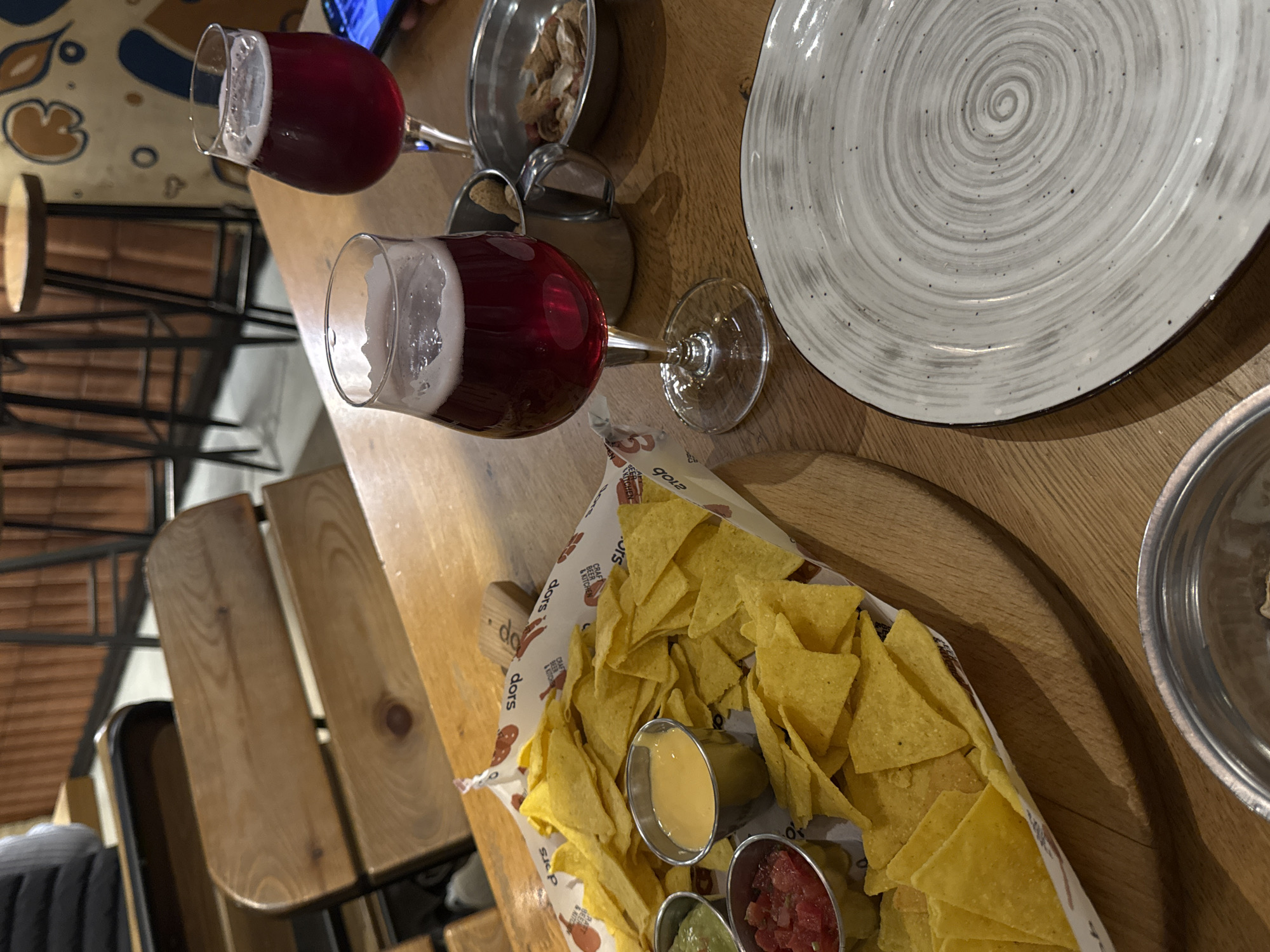 The image depicts a table setting with food and drinks in what appears to be a restaurant or bar. There are two glasses filled with a dark red beverage with foamy heads, likely beer or ale. A round, grey and white patterned plate is present, along with two small metal bowls containing what seem to be snacks or side dishes. In the foreground, a tray filled with tortilla chips and three small containers of dips – likely cheese, salsa, and guacamole – is visible. The table has a wooden surface. Part of a chair is also visible.
The restaurant is "Craft Beer & Kitchen" called "Dors". The "Dors" is located in Yerevan, Armenia.
