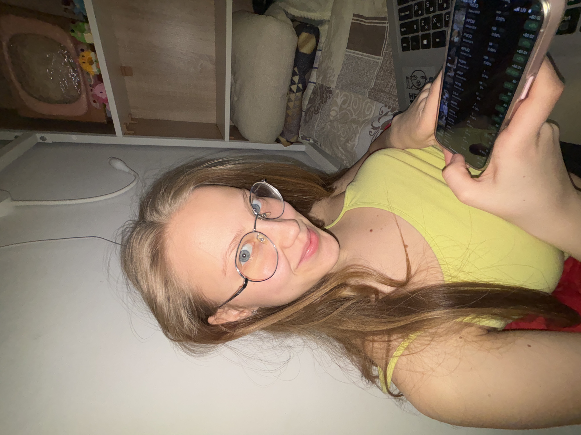 A young woman with long blonde hair and round glasses is lying on her side, looking at her phone. The phone displays stock market data. She is wearing a yellow tank top and appears to be in a room with a desk, keyboard, and various items on shelves. There's no indication to suggest Perm, Russia.