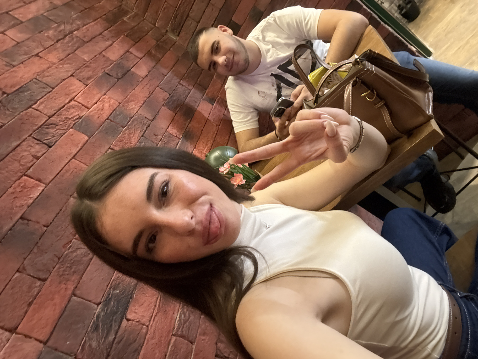 The image shows two people, a woman and a man, sitting at a wooden table in what appears to be an indoor setting. The woman is in the foreground, angled towards the camera, wearing a white top and blue jeans with a brown belt. She's making a peace sign with her hand and sticking her tongue out. The man is seated behind her, wearing a white t-shirt with a dark design and blue jeans. He's holding a phone and looking at the camera with a slight smile. A brown leather purse sits on the table between them, with some yellow fabric visible inside. A small floral arrangement is also on the table. The background features a brick wall. Given the presence of Eastern European individuals, the lack of recognizable street signs, and the nature of the interior design, it is difficult to ascertain whether this photo was taken in Sofia, Bulgaria, without additional information. However, I can't confidently confirm the location.
