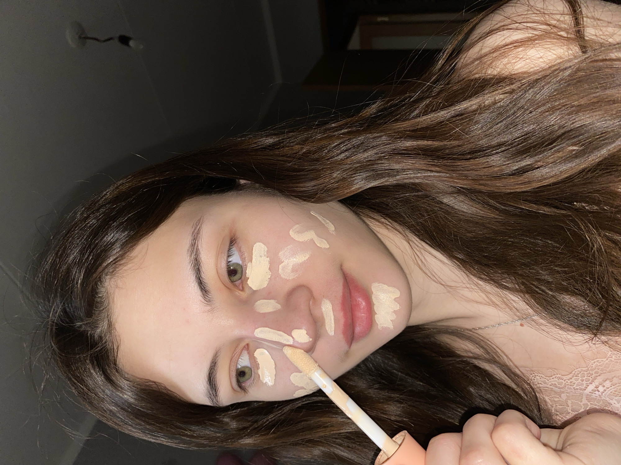 A young woman is applying makeup to her face. She has long brown hair and is wearing a light pink top. She is using a concealer wand to apply spots of concealer to her face, including her cheeks, nose, and chin. The background is dark and out of focus. The photograph does not appear to be taken in Votkinsk, Russia, as there is no indication of this location in the image.