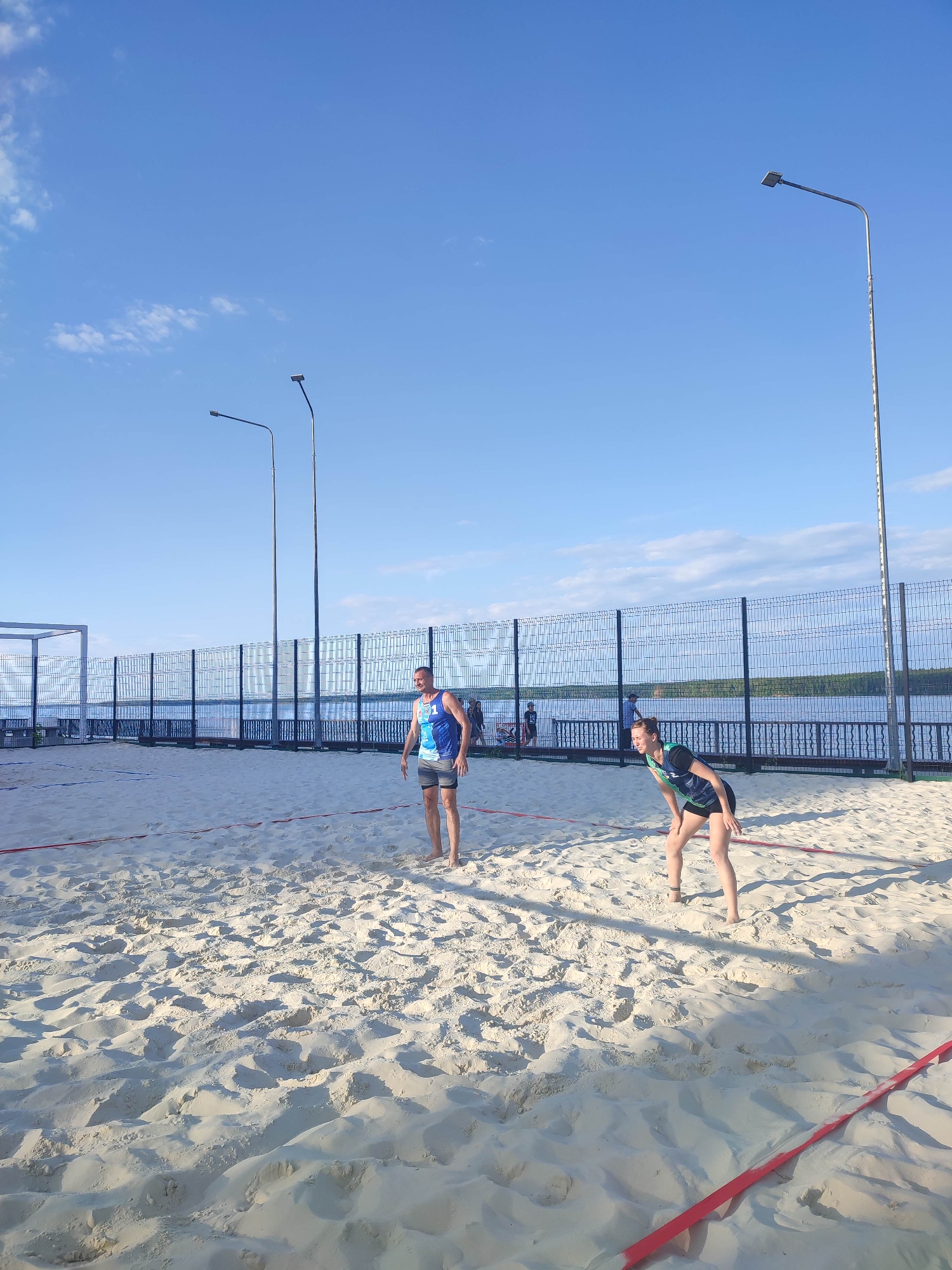 The image depicts a beach volleyball court on a sunny day. Two individuals are visible on the sandy court, actively engaged in a game. A male player, wearing a blue and white tank top and grey shorts, stands on the left side of the court, facing the net. On the right side, a female player, dressed in a black and green tank top and black shorts, is in a defensive stance, ready to receive the ball.
Behind the court, a tall wire fence encloses the area, beyond which a body of water, likely a lake or river, is visible. The water reflects the clear blue sky, which has a few scattered white clouds. Two tall lampposts are positioned behind the fence, suggesting the court might be used in the evening or at night. In the distance, a white structure, possibly a pavilion or shelter, can be seen on the left side of the frame. The sand on the court is well-raked, with visible lines marking the boundaries of the court. Red tape is laid out along the sidelines. The overall impression is a bright, pleasant day for outdoor sports.
