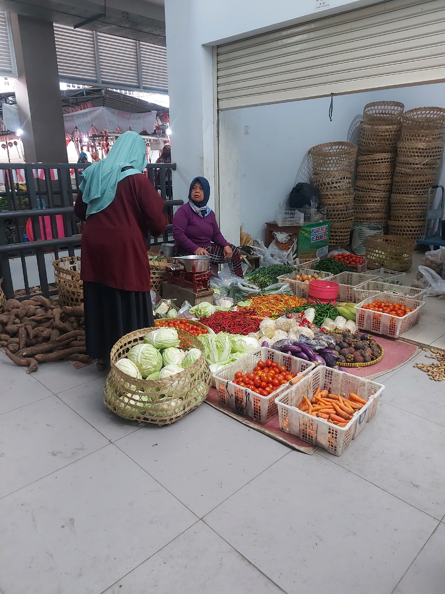 The image shows an outdoor market scene in Godean, Indonesia. Two women are present. One woman, wearing a turquoise headscarf and a maroon long-sleeved shirt and dark skirt, stands facing away from the camera, seemingly interacting with a vendor or customer. The second woman, wearing a dark blue headscarf and a purple top, sits behind a stall laden with various fresh produce, including cabbage, tomatoes, chilies, eggplants, mangosteens, and carrots. She appears to be a vendor, with a red scale visible on her stall. The market environment is characterized by a tiled floor, a covered walkway or roof structure above, and stacked woven baskets in the background. The scene suggests a typical bustling local market day.