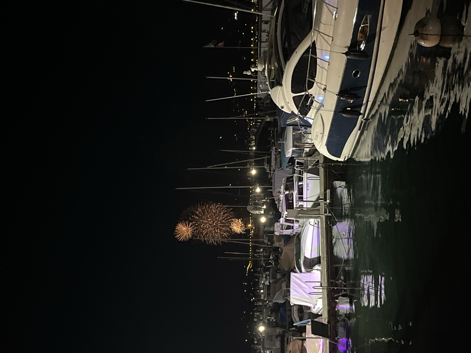 This image depicts a nighttime scene in the city of Geneva, Switzerland, likely at a marina during a fireworks display. In the foreground, several boats are docked. A large, white yacht with blue trim is prominently featured on the right, its hull reflecting in the dark water. Several smaller boats, some covered with white tarps, are moored to the left.
In the background, fireworks are bursting in the night sky, casting a warm glow. The lights of the city can be seen as a distant, blurred line along the horizon. The overall atmosphere is festive and celebratory. The water is calm, with reflections of the fireworks and boat lights creating an abstract pattern on its surface. There is no indication of people actively engaged in any specific actions, but their presence is implied by the activity of the fireworks and the illuminated city. The environment is a waterfront setting, characteristic of Geneva's lakeside location.
