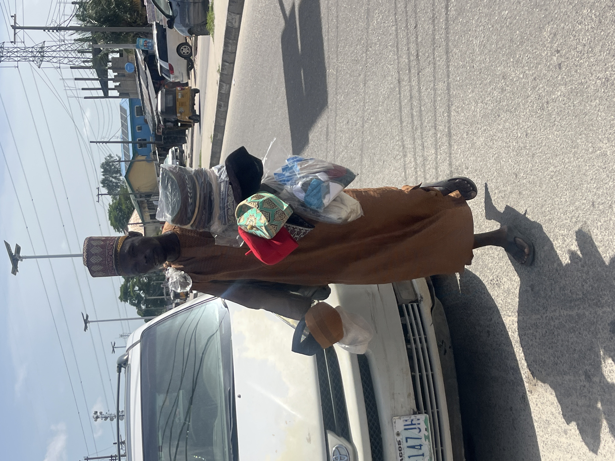 An image taken in the city of Ado, Nigeria, shows an adult male street vendor wearing a brown garment and a patterned brown cap. He is standing behind the front of a white car, likely using it as a display for his wares, which include various types of hats and caps piled on his back and placed on the car's hood. The car has a license plate that reads "LAGOS 147 JH". The surrounding environment includes a paved road, power lines, and buildings in the background.