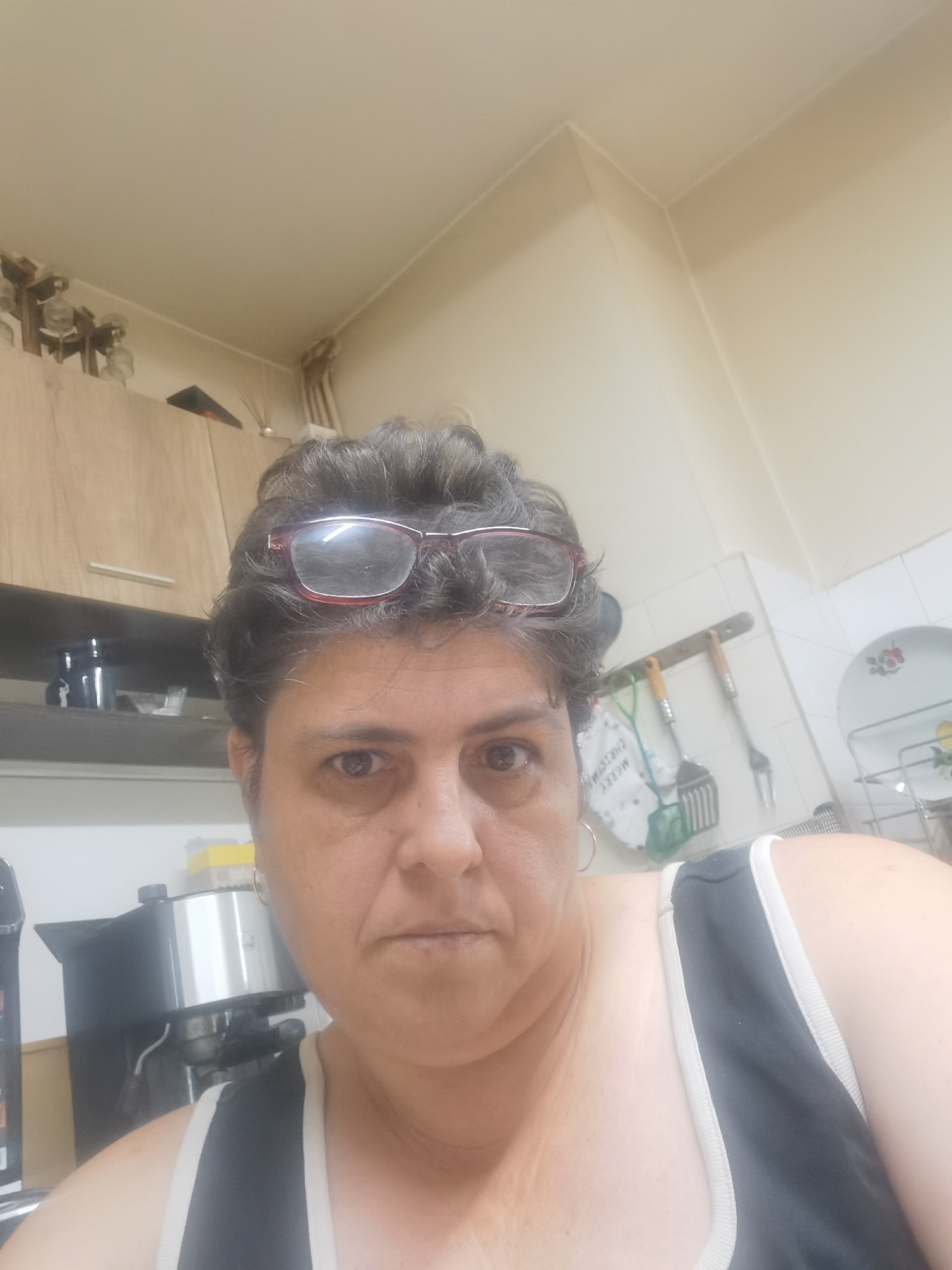 A medium shot selfie shows a middle-aged woman with graying, curly hair and glasses perched on her forehead. She is wearing a black tank top with white trim. Her expression is neutral, and she is looking directly at the camera. The background appears to be a kitchen, with cabinets, a coffee maker, and utensils visible. The lighting is bright, suggesting it might be daytime. The image does not provide any specific information about the location in the city of Lomets, Bulgaria.
