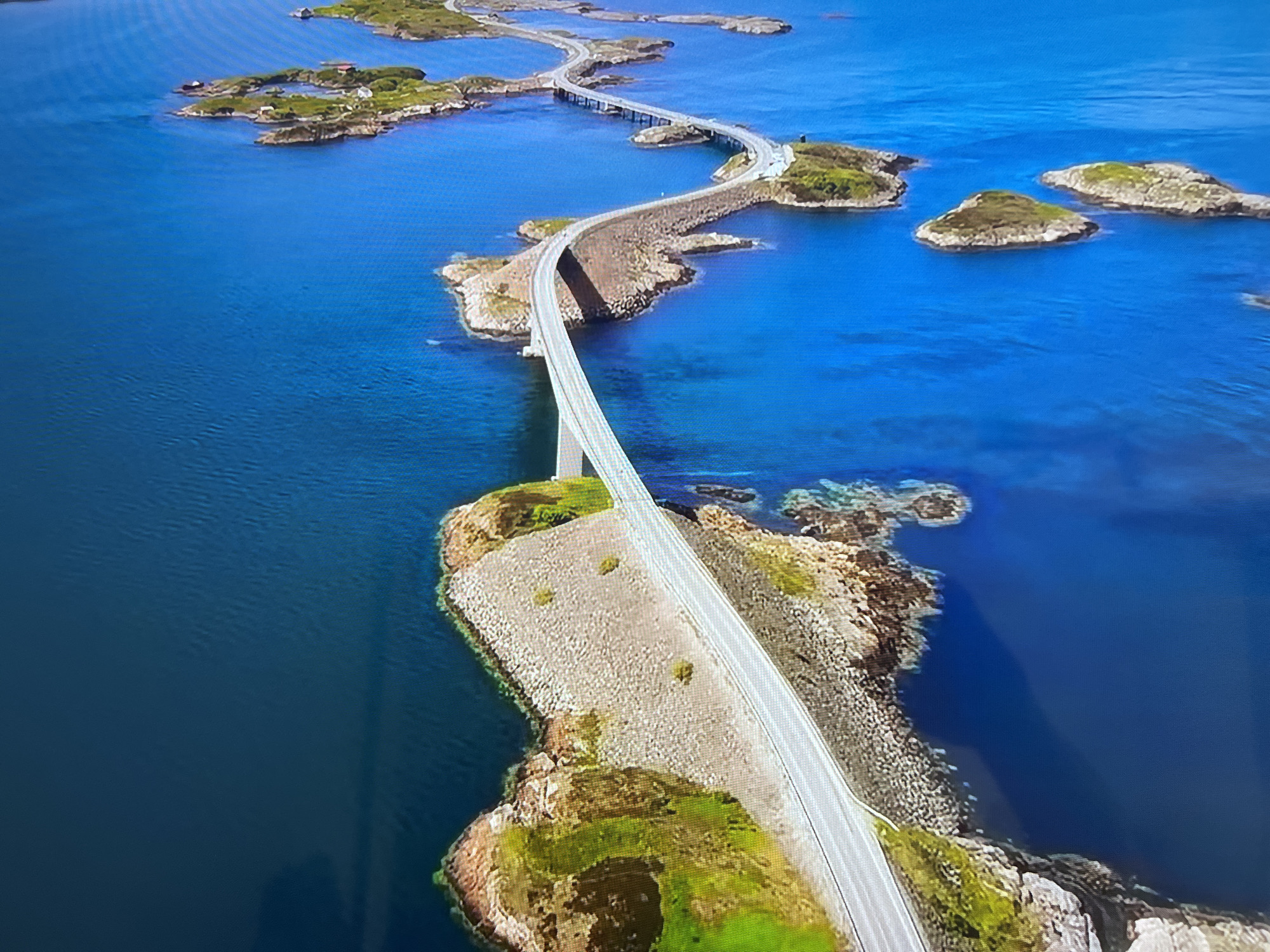 The image depicts the Atlantic Road in Norway. The road is a winding sequence of bridges and causeways connecting islands and skerries. The water is a deep blue, and the islands are rocky with patches of green vegetation. There is no evidence of the location being in Basingstoke and Deane, United Kingdom.