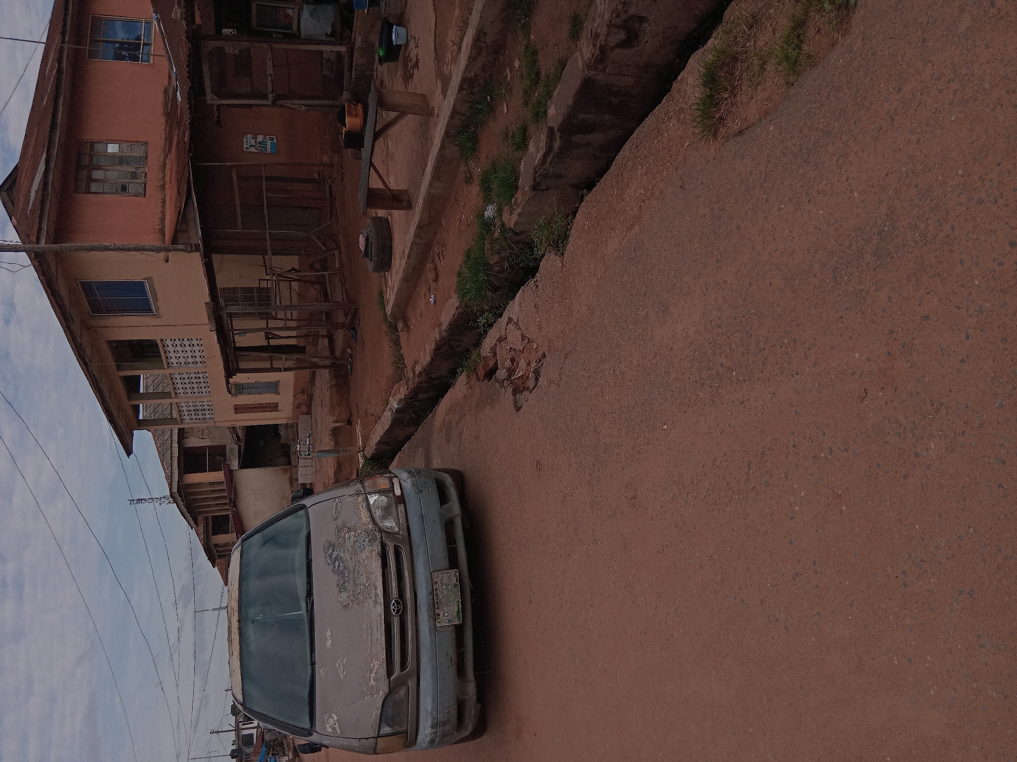 The image depicts a street scene in Shagamu, Nigeria. An old, faded silver Toyota vehicle is parked on the side of a reddish-brown road. The vehicle has a green and white license plate. Adjacent to the road is a concrete drainage ditch with some vegetation growing in it. Buildings line the street; they appear to be two-story structures made of brick and wood, with weathered paint. Overhead power lines are visible against a pale sky. The overall scene suggests a low-income residential area.