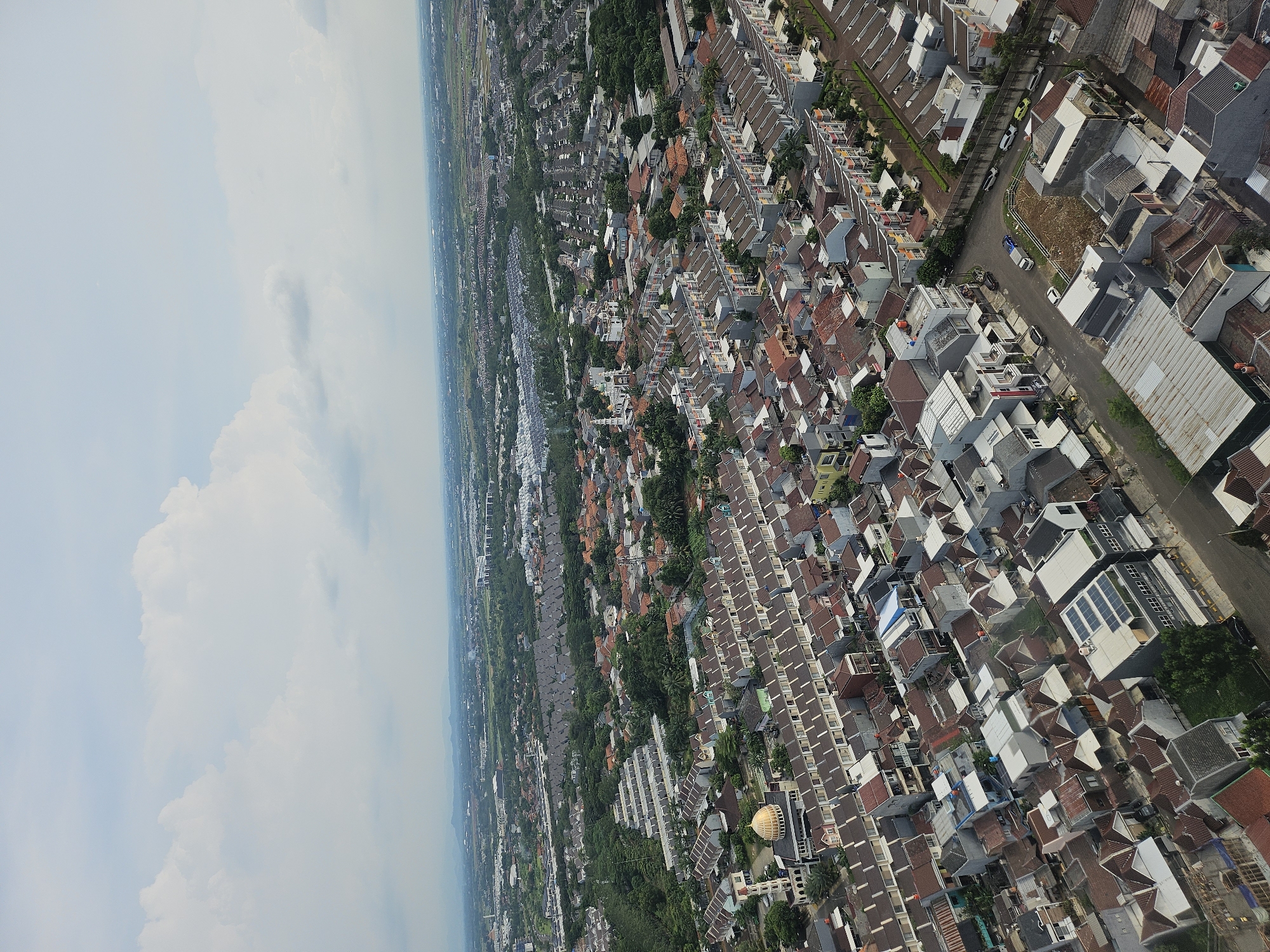 The image presents an aerial view of a residential area in Medang, Indonesia. The architecture is dense, consisting primarily of single-family homes with varying roof colors and styles, predominantly brown and grey. A mix of modern and traditional construction is visible. Streets are narrow and lined with sparse vegetation. In the background, there are rice fields, and the sky is partially covered with clouds.
