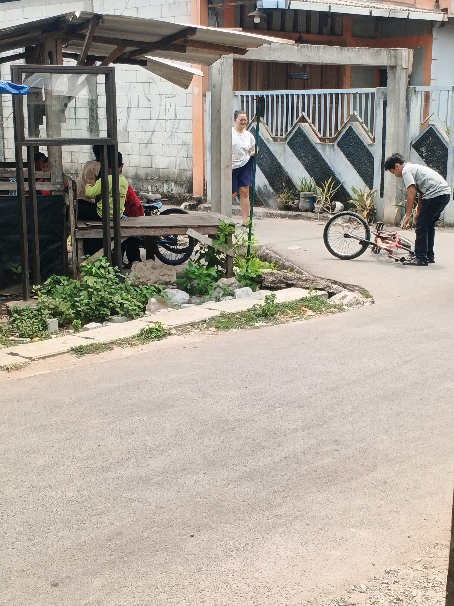 In Kadu, Indonesia, a young man is seen on the right side of the image, bending over to attend to a bicycle that has fallen on its side in the street. He is wearing a gray t-shirt and dark pants. Behind him, a woman with glasses and wearing a white patterned shirt and blue shorts stands near a decorative fence. To the left, a simple wooden structure, possibly a shelter or shop, is visible with three individuals seated within. One person wears a light brown shirt, another a bright yellow shirt, and the third is partially obscured by the red clothing of another person. The ground appears to be a paved road and sidewalk area, with some overgrown vegetation and scattered debris. The background shows a building with a corrugated metal roof and a brick wall.