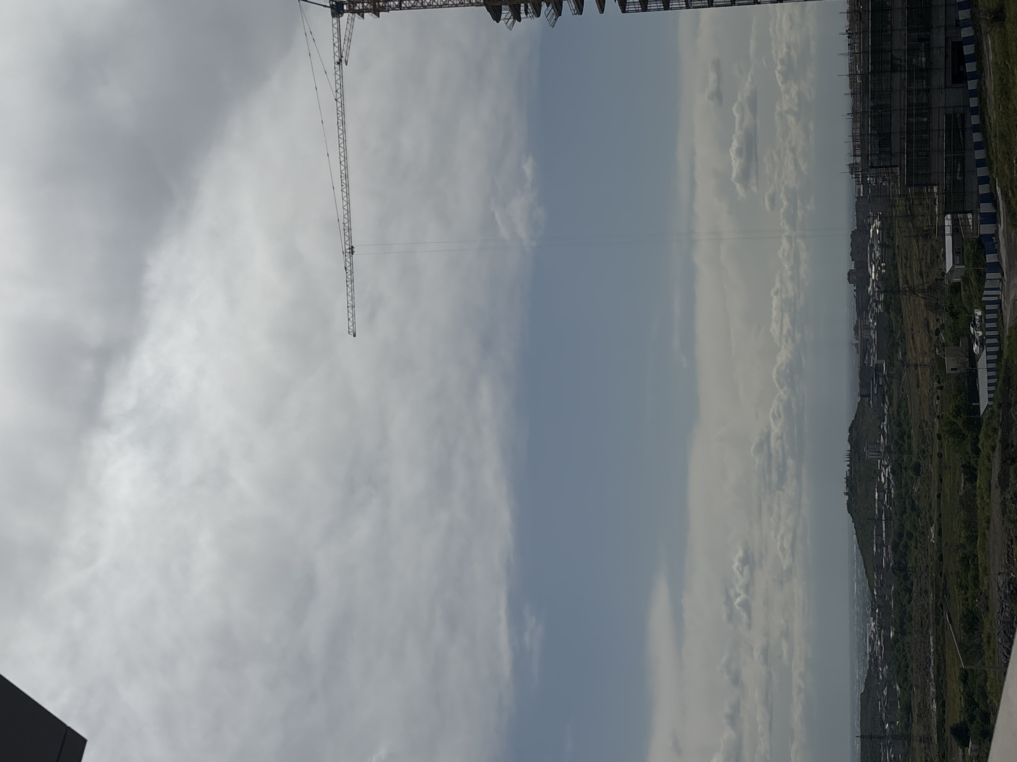 The image depicts an overcast sky above Yerevan, Armenia. A construction crane is visible on the left, towering over a construction site with partially built structures. The skyline in the distance reveals an urban landscape with buildings nestled amidst green hills. The overall environment suggests ongoing construction activity within the city.