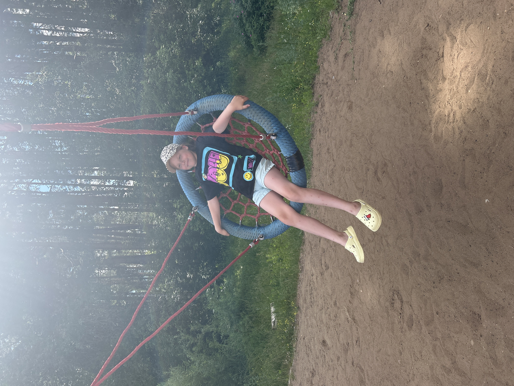 A young girl is sitting on a round, blue rope swing with red ropes suspending it. She is wearing a black t-shirt with a colorful graphic, light blue shorts, a leopard-print baseball cap, and yellow clogs. She is smiling and looking slightly to her left. The swing is in a sandy area with a grassy background and tall trees behind it, suggesting an outdoor park or playground setting. The lighting indicates it is daytime, possibly with some dappled sunlight. There is no information in the image or provided context to determine the specific location within the city of Chaikovsky, Russia.