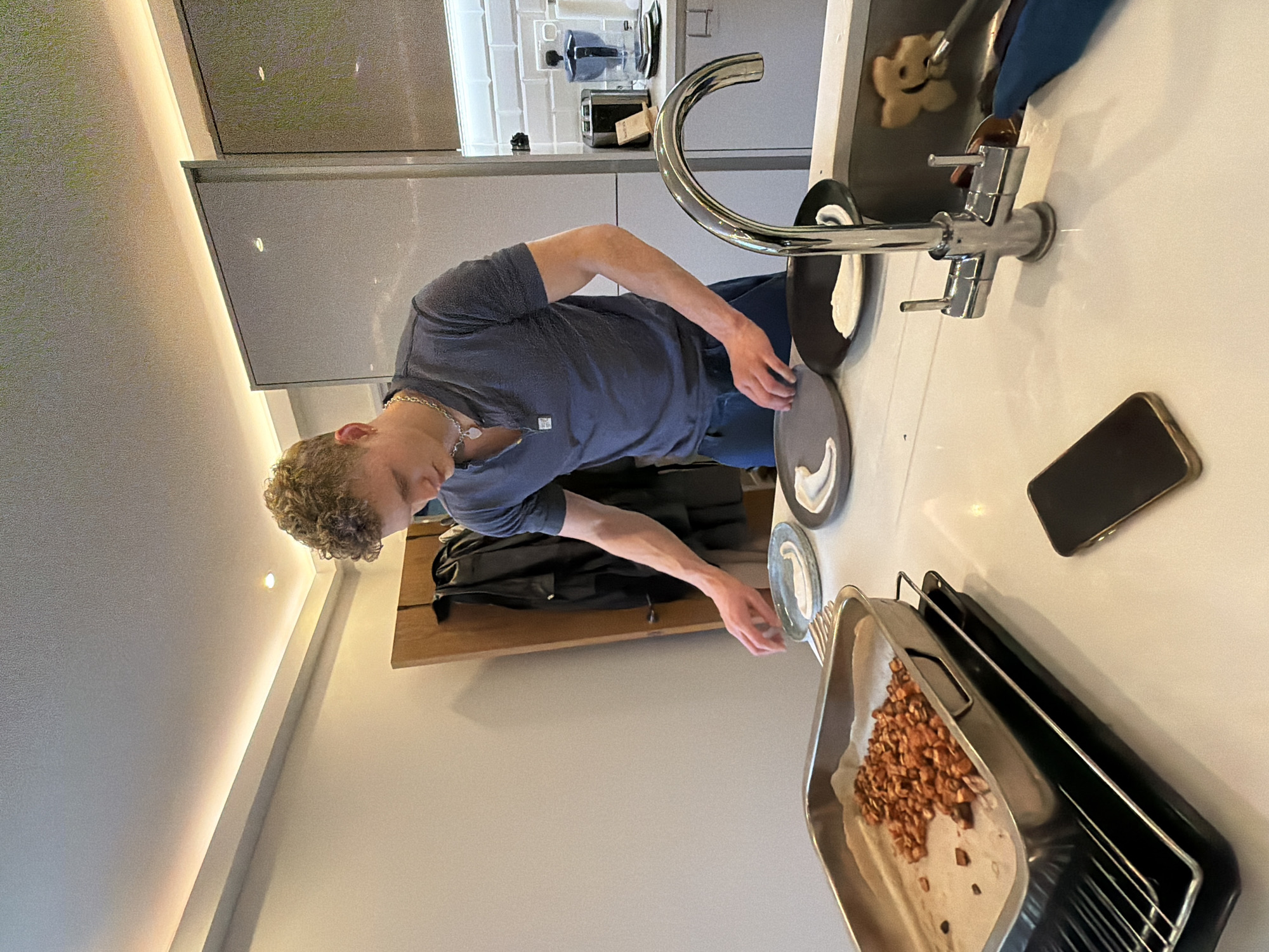 The photograph depicts a young man in a domestic kitchen setting. He is fair-skinned with curly blonde hair, wearing a short-sleeved blue t-shirt and a silver chain necklace. The man is leaning over a kitchen countertop, appearing to be engaged in washing dishes. Several plates are stacked in or near the kitchen sink. A baking tray filled with what appears to be cooked or toasted nuts is located on the right side of the frame. A mobile phone rests on the countertop nearby. A black jacket hangs on a wooden wall mounted fixture behind the man.
The kitchen has modern, minimalist decor. The cabinets are a light grey or off-white, and the countertop is a light-colored composite material. The appliances include a silver faucet. The scene is well-lit, likely from overhead lighting. There is nothing in the image to suggest that it is specifically located in the city of London, or the United Kingdom.