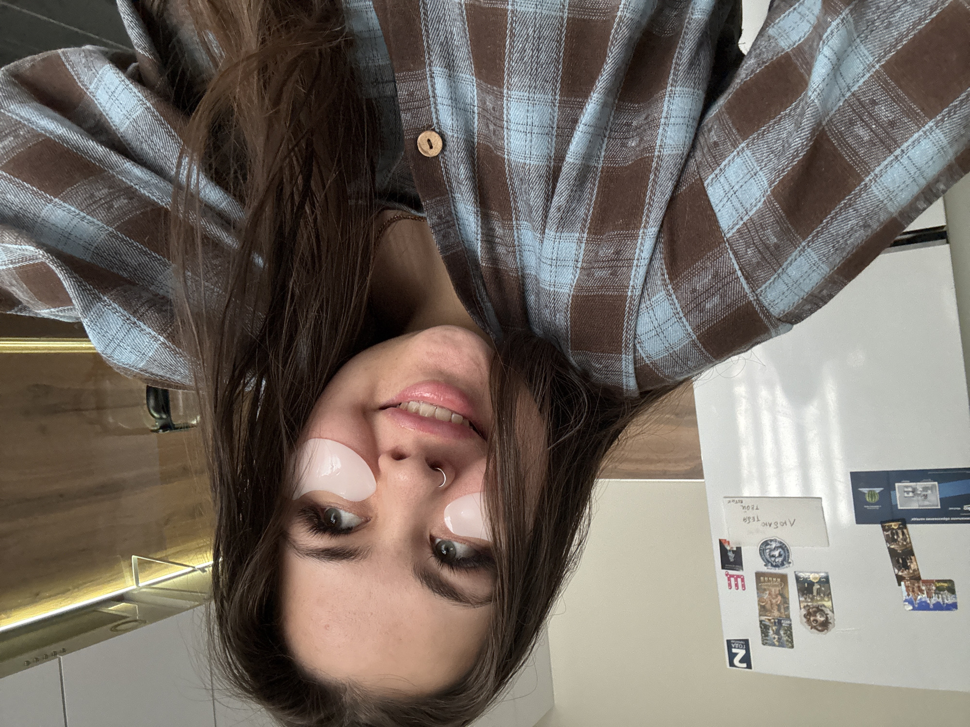 An upside-down selfie shows a young woman indoors, likely in a home kitchen. She has long, dark brown hair and is wearing a brown and light blue plaid shirt with a visible wooden button. She has pinkish, gel-like under-eye patches applied under both eyes and a silver hoop nose ring. The woman is smiling slightly, looking directly at the camera, suggesting a relaxed and positive mood while engaging in a self-care routine.
To her left, a modern kitchen counter with an under-cabinet light fixture is visible. To her right, a white refrigerator is covered with various magnets and notes. A prominent handwritten note reads "ЛЮБЛЮ ТЕБЯ ТЕТЯ" (I LOVE YOU AUNT) with "ТЕТЯК" below it. Other magnets include one with "MUMIY" (likely a reference to the Russian band Mumiy Troll), one featuring a stylized logo or crest, and another with "ТОЛЯ" (TOLYA) and the number "2", alongside several other decorative magnets and small pictures. The image, taken with an inverted perspective, captures a personal moment within a domestic setting, located in Bugry, Russia.