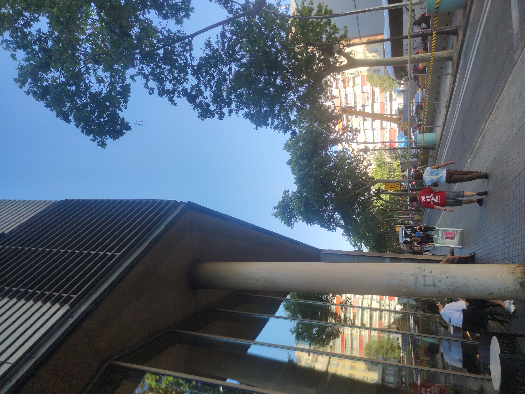 A street-level view in Barcelona, Spain features a mix of architecture and pedestrian activity. A modern building with a slatted exterior and large glass windows is in the foreground, supported by a cylindrical concrete column. Reflections in the glass show the street and surrounding buildings. In the background, a row of tall apartment buildings stands along the street. Trees line the road, providing partial shade. People are walking along the sidewalk, and a roadside barrier runs parallel to the street. A small sign or advertisement is on the sidewalk near the people. The sky is blue and visible through the trees.