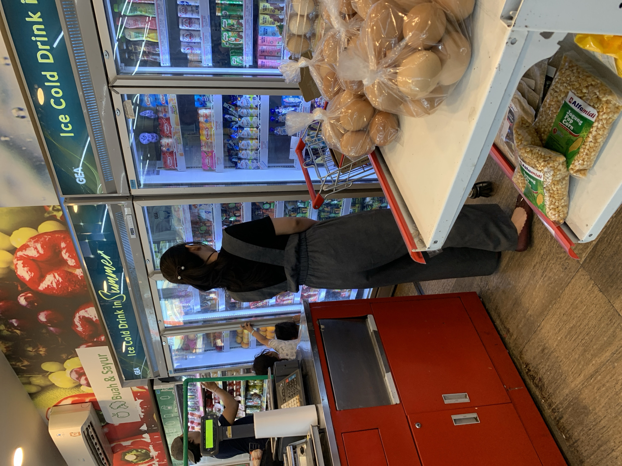 This is a medium-angle, eye-level shot of a retail store with a cashier, customers, and shelves stocked with food and drinks. A woman in a black top and grey pants is standing in front of a refrigerator, reaching for something inside. To her right, a cashier is operating a point-of-sale system. In the foreground, a red cabinet is visible, with bags of popcorn on a shelf above it. The store appears to be well-lit and stocked, with products ranging from drinks and produce to packaged goods. Visible text includes brand names like "GEA," "Alfamidi," "Yakult," and "MILO," along with promotional signs in Indonesian, such as "Ice Cold Drink in Summer" and "Buah & Sayur" (Fruits & Vegetables). The environment suggests an indoor grocery or convenience store setting, possibly in Makassar, Indonesia, given the language on the signs. The overall mood is neutral, reflecting a typical retail transaction. There are no clear indicators of the time of day or weather outside.