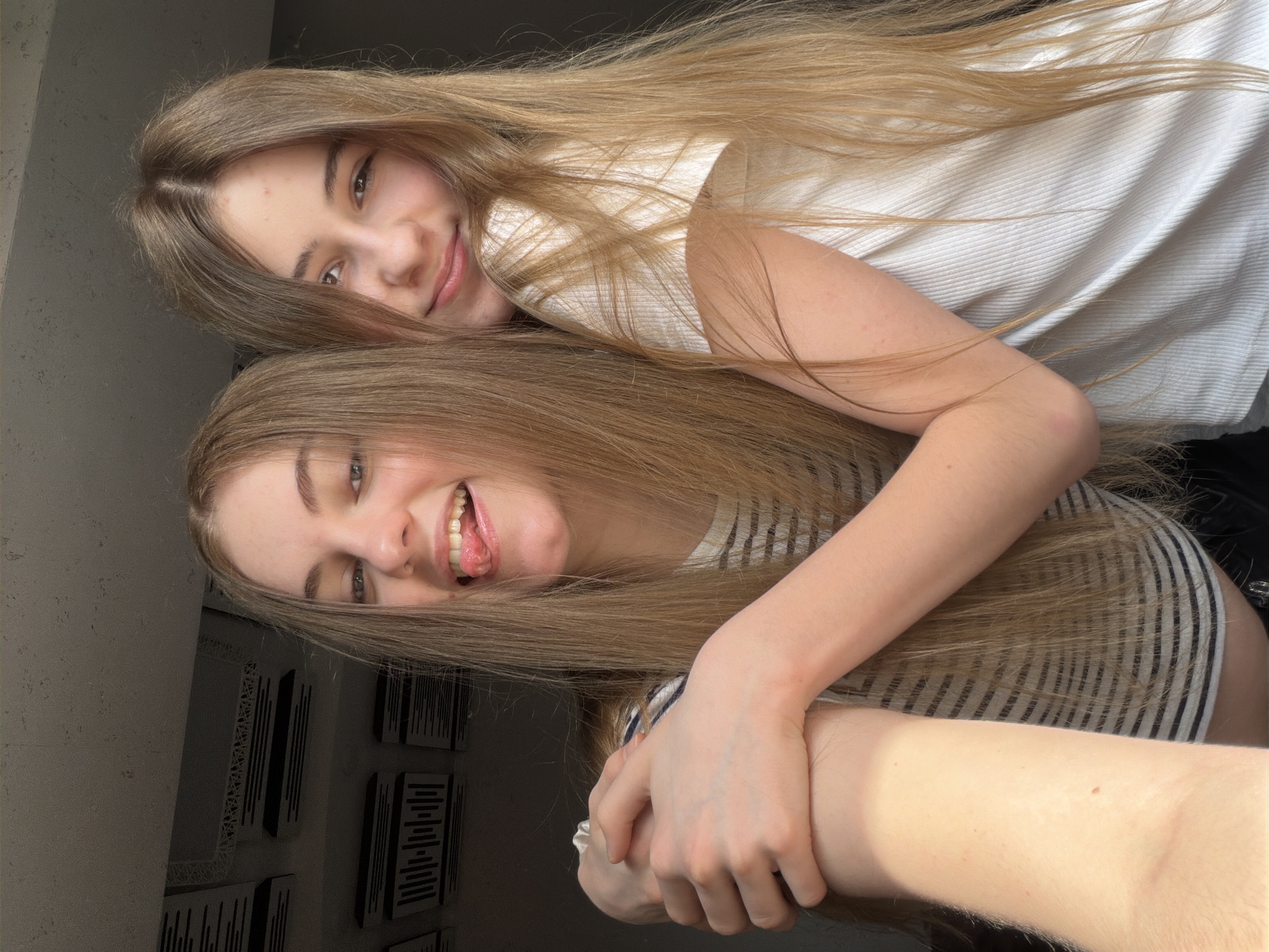 Two young women, likely teenagers, are posing for a selfie indoors. The woman on the left has her tongue sticking out playfully, while the woman on the right smiles at the camera. Both have long, blonde hair. They are wearing casual clothing; the woman on the left is in a striped shirt, and the woman on the right is in a white ribbed t-shirt. The background appears to be a room with acoustic paneling on the ceiling. The lighting suggests it might be daytime, with sunlight coming from a window. The scene is likely a personal residence or a casual studio space in Dnipro, Ukraine.