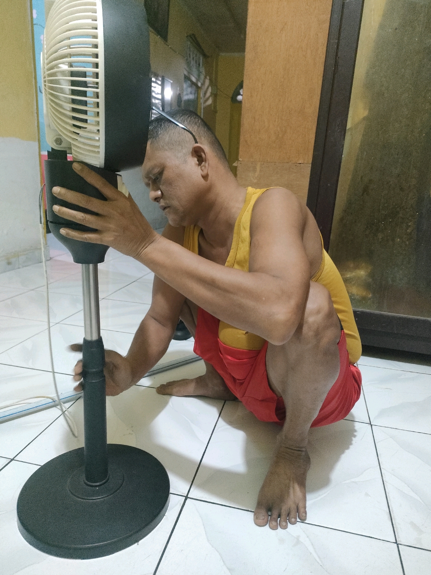 An adult male is shown kneeling on a tiled floor, interacting with the stand of a white and black standing fan. The man is wearing a yellow tank top and red shorts. He has sunglasses perched on his head. The background includes a wall and what appears to be a doorway or window with a reflective surface. The environment suggests an indoor setting, possibly a residential or commercial space in Pekanbaru, Indonesia, indicated by the visible details of the room and the overall aesthetic. The man's hands are positioned on the fan's stand, suggesting he is adjusting or maintaining it. The lighting is moderate, casting some shadows on the floor and the man's body.