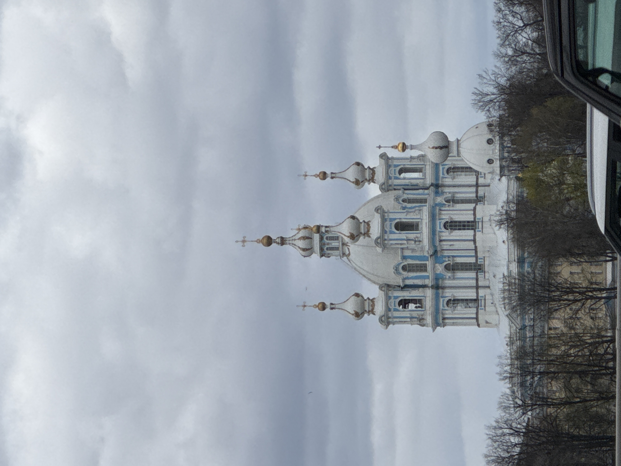 The image shows the Smolny Cathedral in Saint Petersburg, Russia, from a ground-level perspective, possibly taken from inside a vehicle. The church's architecture features white walls with blue accents, multiple domes with golden spires, and visible windows. The sky is overcast, contributing to the muted lighting. In the foreground, part of a vehicle is visible, suggesting the photograph was taken from inside. Bare trees surround the cathedral, indicating a time of year when the leaves are off the trees. Another building is partially visible behind the trees to the right of the cathedral.