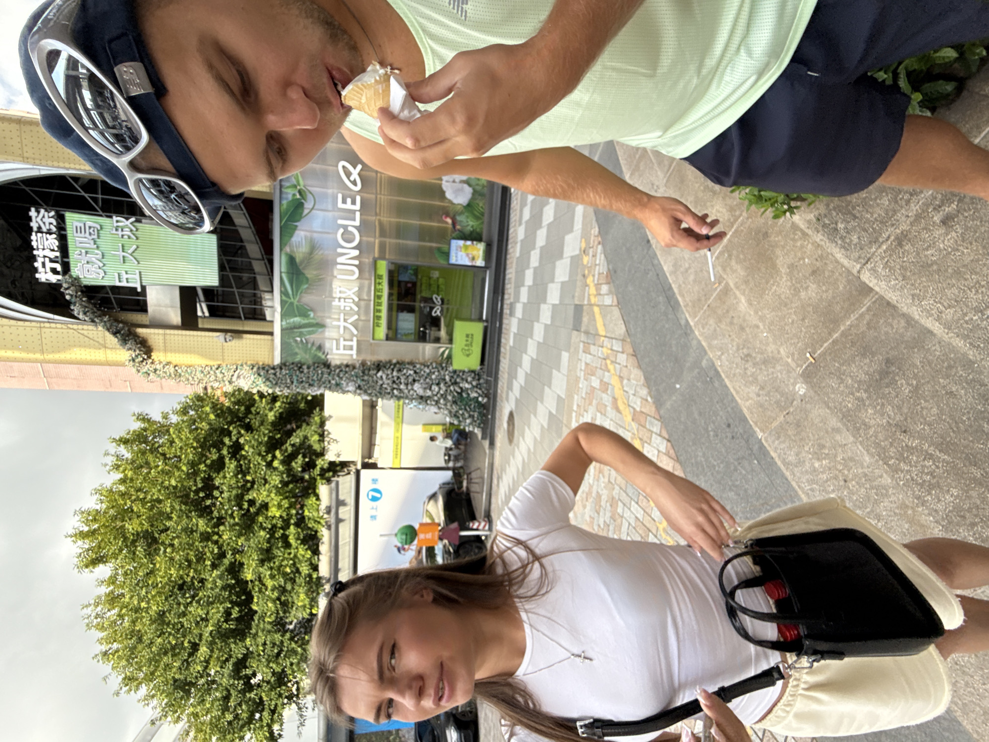 In Haizhu District, China, a young woman with blonde hair tied back stands to the left, wearing a white t-shirt, a cross necklace, and a cream-colored skirt. She carries a small black purse. To the right, a man wearing a light green tank top, navy shorts, and a blue baseball cap with sunglasses on top is eating an ice cream cone. He holds a cigarette in his right hand. The background shows a building with Chinese signage, a large green tree, and a paved sidewalk with patterned tiles.