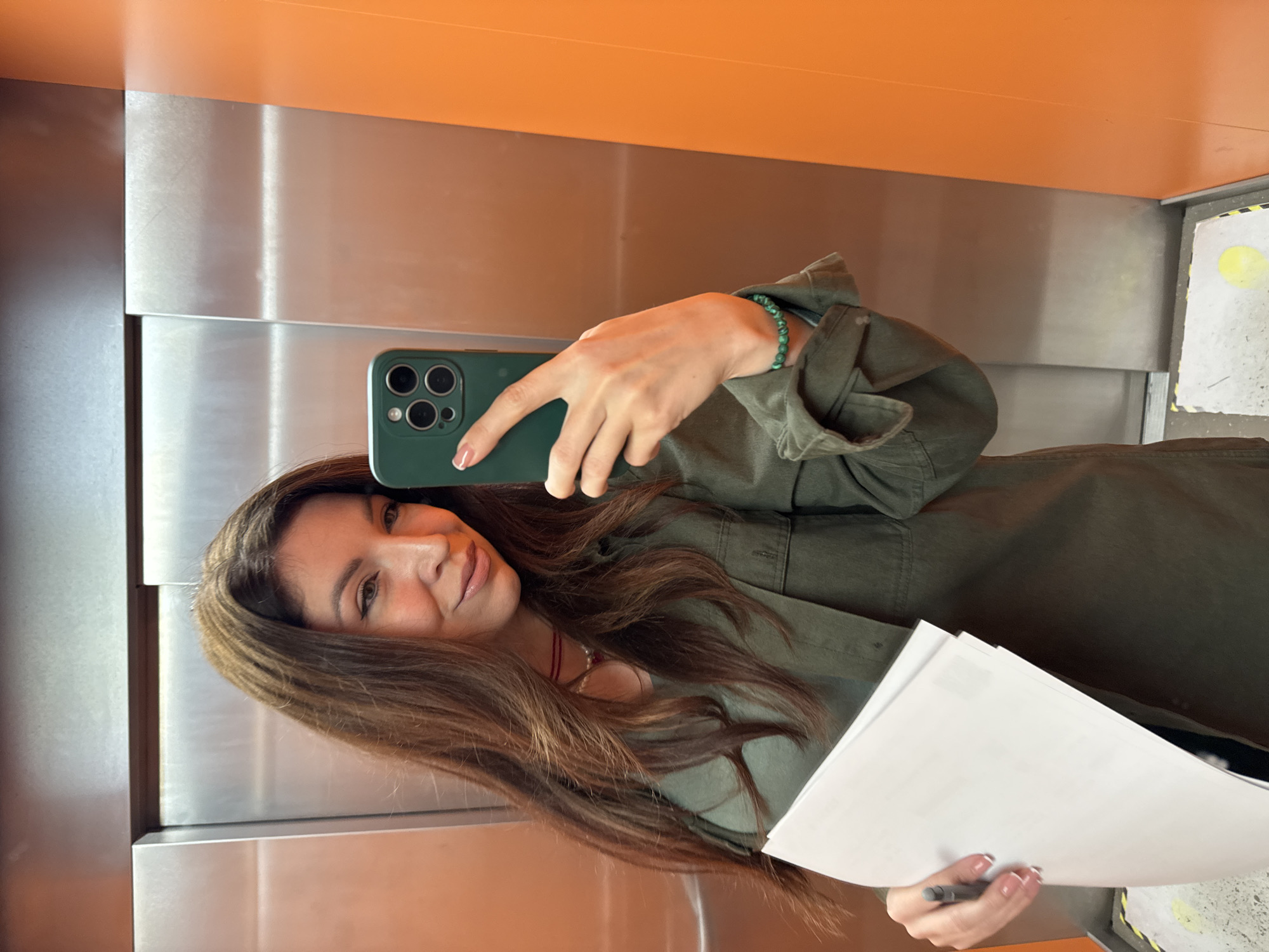 A woman with brown hair and an olive-green shirt is taking a selfie in an elevator. She holds a phone in her right hand and some white papers in her left. The elevator has metal walls and an orange ceiling. A section of the floor in the background has what appear to be yellow marks on the paneling. This image was taken inside an elevator, which is not a sufficient clue to geolocate to Kryvyi Rih.