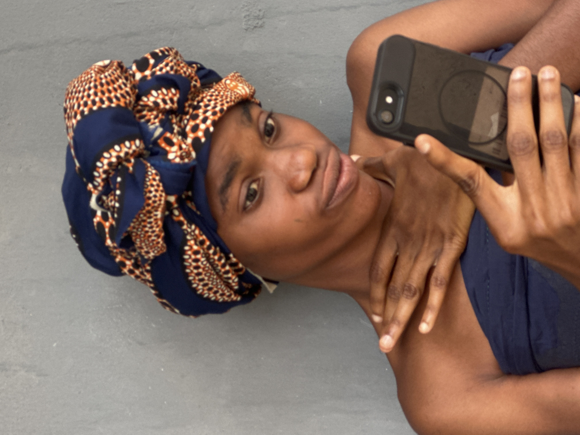 A young Black woman is taking a selfie with her phone in front of a grey, textured wall. She is wearing a colorful headwrap with a dark blue base and an orange and white patterned design. She has her left hand resting on her chest and her right hand holding a black smartphone. Her head is slightly tilted, and she appears to be posing for the photo. The background wall suggests an outdoor setting. The image does not provide any specific indicators to pinpoint the location to Badagry, Nigeria.