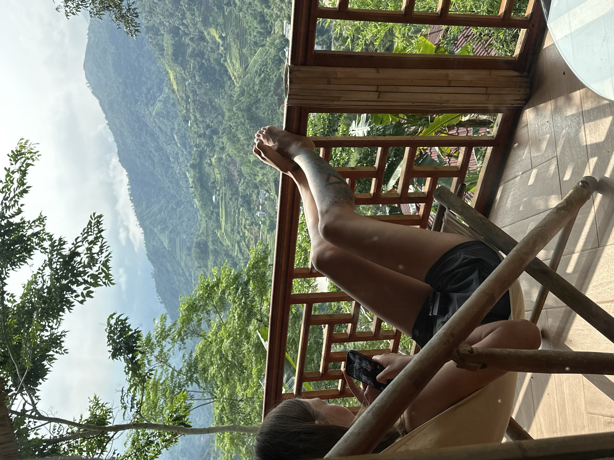 The image depicts a person relaxing on a wooden balcony, overlooking a vast, lush green mountainous landscape in Thông Nguyên Commune, Vietnam. The main subject, appearing to be a young woman, is lounging in a light-colored, possibly fabric and bamboo, chair. Her feet are propped up on the dark wooden railing of the balcony, and she has tattoos visible on her legs. She is wearing dark shorts and is holding a black smartphone with both hands, seemingly engrossed in its contents.
The scene is set outdoors during daytime, with bright natural light casting shadows on the wooden floor and railings of the balcony, suggesting a sunny or partly cloudy sky. The background features rolling mountains covered in dense vegetation, with some terraced fields visible on the slopes, characteristic of agricultural practices in mountainous regions. Clouds partially obscure the peaks in the distance. To the left, leafy trees frame the view, enhancing the natural setting.
The overall atmosphere is one of leisure and contemplation, combining modern technology with a serene natural environment. No visible text can be discerned on the smartphone screen or any other part of the image.
