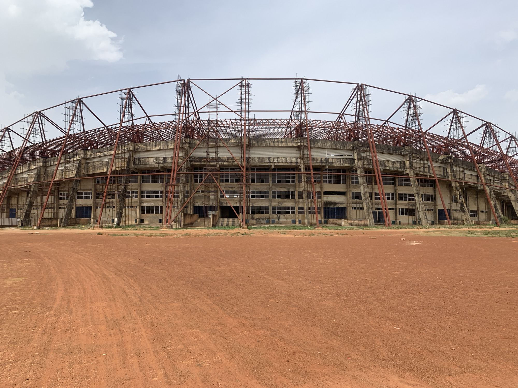 The image depicts a large stadium under construction or renovation in Rafin Par, Nigeria. The stadium's exterior is predominantly concrete, with visible window rows and a steel framework surrounding the structure. The framework suggests ongoing construction or repair work. The foreground is a reddish-brown dirt surface, likely a field or open space adjacent to the stadium. The sky is overcast with scattered clouds. There are no people visible in the frame.