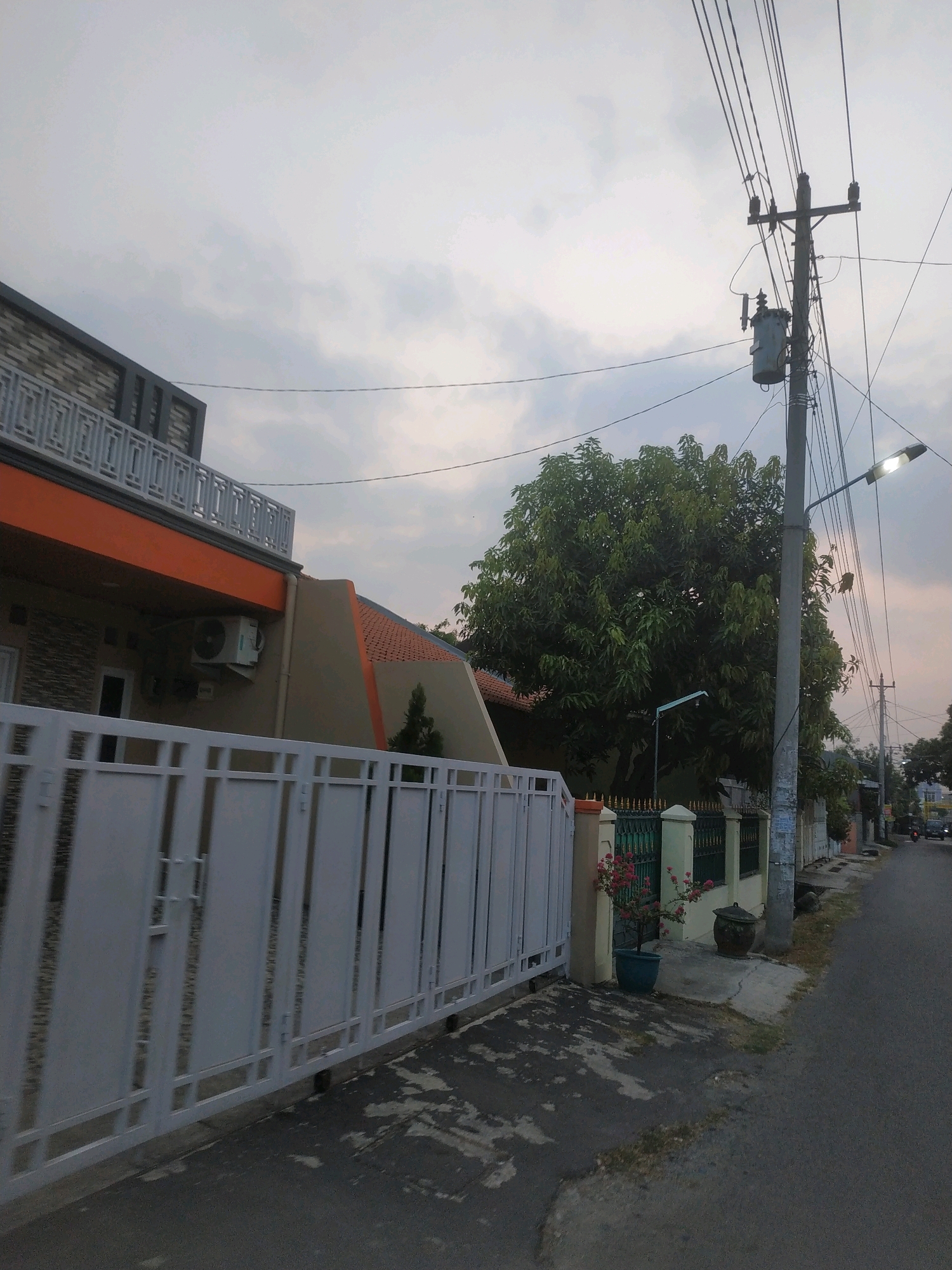 The image captures a street view in Tegal, Indonesia, likely during dusk or an overcast day. A modern house with a white fence and an orange accent on the second story is visible on the left. An air conditioning unit is mounted on the wall of the house. To the right of the house, a street stretches into the distance, lined with houses, a tall utility pole with a transformer, and a street lamp that is illuminated. A large, leafy tree stands prominently in the middle ground. A pink flowering plant in a pot and a dark, round urn are situated near the fence. The road surface appears worn and cracked. The sky is filled with scattered clouds. In the distance, a vehicle can be seen on the road. The overall scene depicts a residential area in an Indonesian city.