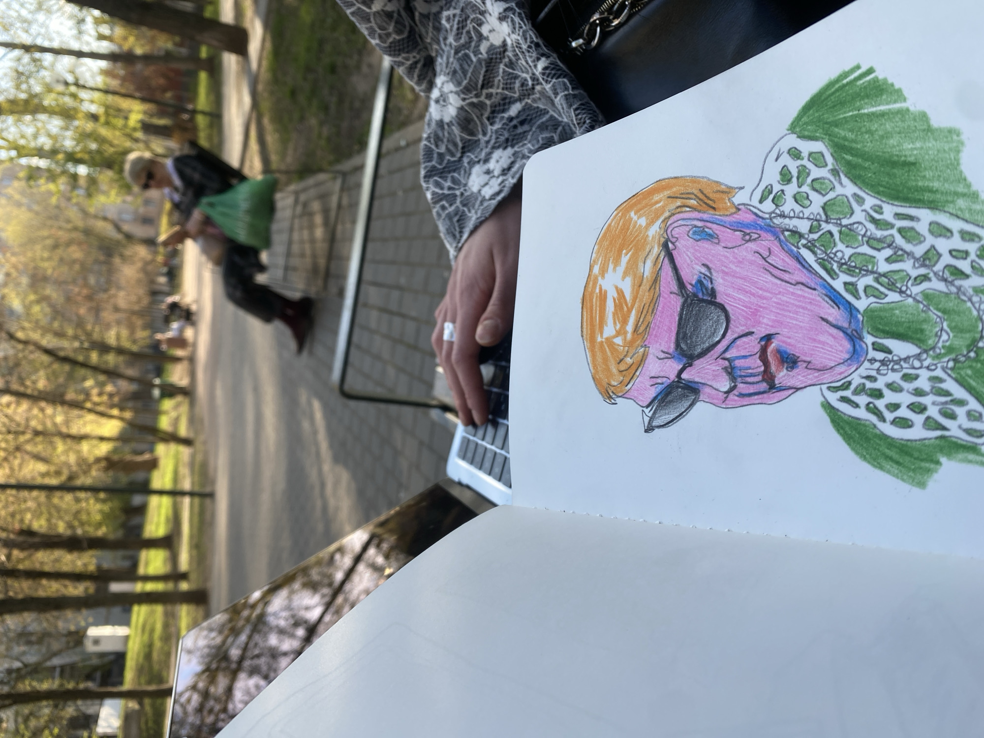 The image shows a person sitting outside in a park. The location is most likely in a park in Moscow, Russia based on the style of architecture and foliage. The person is sitting at a table or bench with a laptop and a sketchbook. In the sketchbook, there is a drawing of a person with pink skin, orange hair, an eye patch, and a green patterned shawl or cape. In the background, there is another person sitting on a bench with a green bag. There are trees and a walkway in the background, suggesting the park setting. The overall impression is of someone relaxing and sketching in a park setting.
