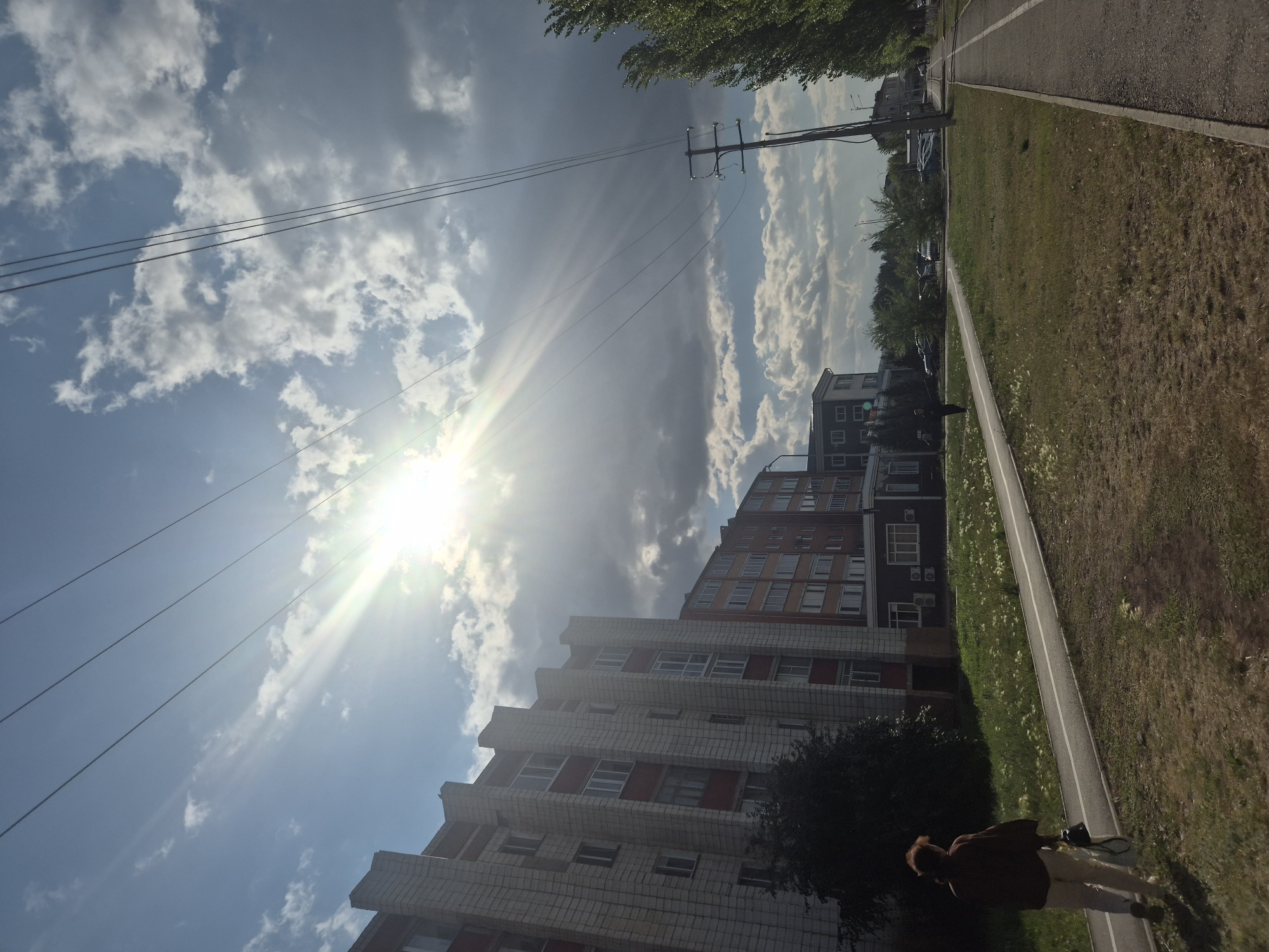 A low-angle shot captures a sun-drenched urban scene. In the foreground, a paved path, possibly a bicycle lane, runs diagonally from the bottom left to the top right of the frame. A person walks away from the camera on this path. To the left of the path are buildings, the closest one being a multi-story apartment building with a light-colored brick facade accented with darker red. The sun is bright and positioned high in the sky, casting long rays that streak through dramatic clouds. Power lines stretch across the sky, supported by a wooden pole visible in the mid-ground. Beyond the initial apartment building, other structures are visible, including a narrower, darker building. The environment appears to be a park-like area with green grass and scattered trees. The scene suggests a typical day in an urban residential area.
