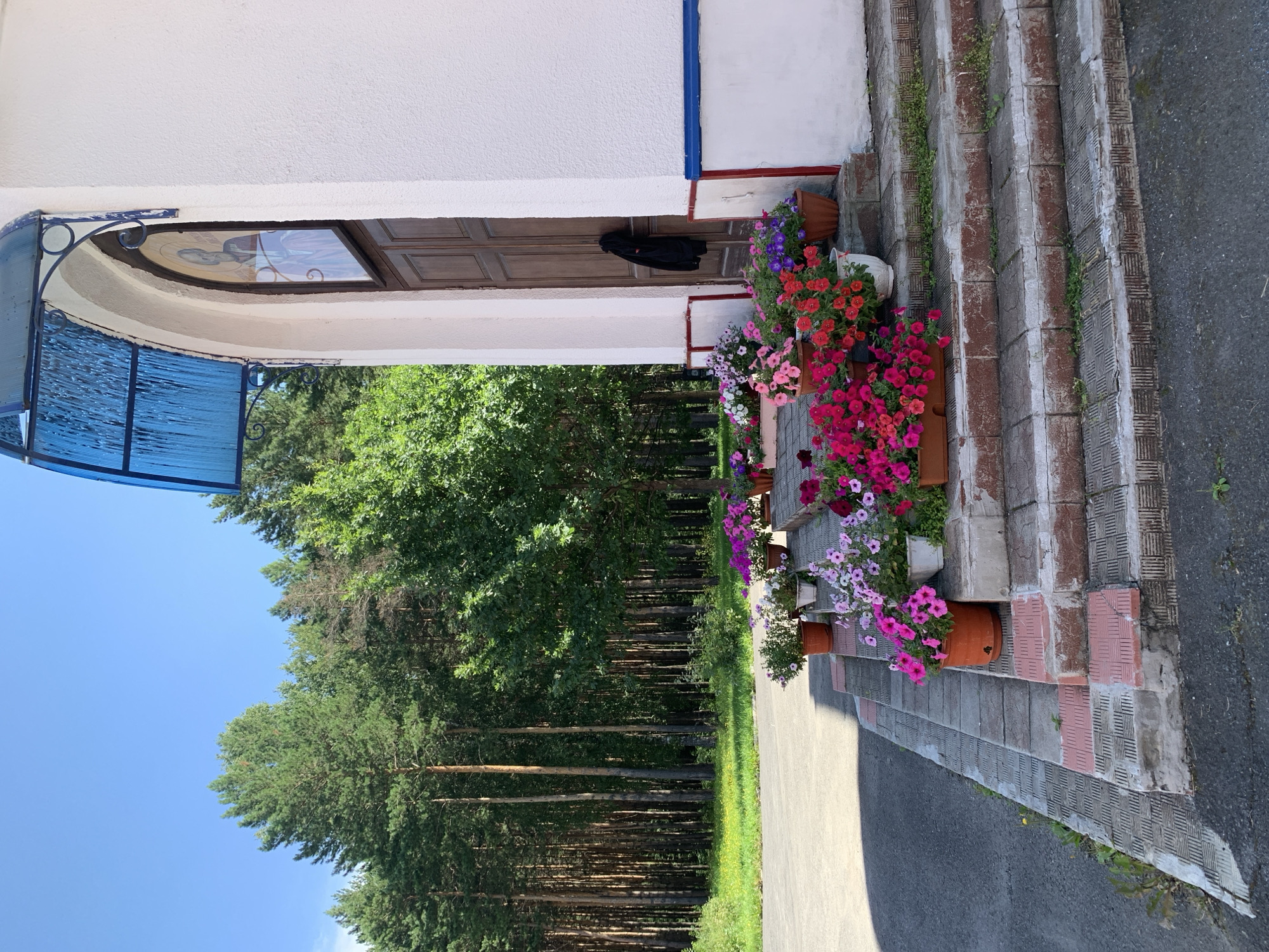 The image shows the entrance of a building, likely a church, with a white facade and a wooden door. The entrance is adorned with numerous potted flowers, predominantly petunias, in various shades of pink, red, and purple. The building is situated in a natural setting with a dense forest of pine trees in the background. The ground in front of the entrance is paved with stone tiles, and a road is visible to the left. The sky is clear and blue. The overall impression is one of a peaceful and well-maintained religious site surrounded by nature. This scene is consistent with the aesthetic of many churches and chapels found in rural or suburban areas of Prokopyevsk, Russia, a city known for its proximity to natural landscapes.