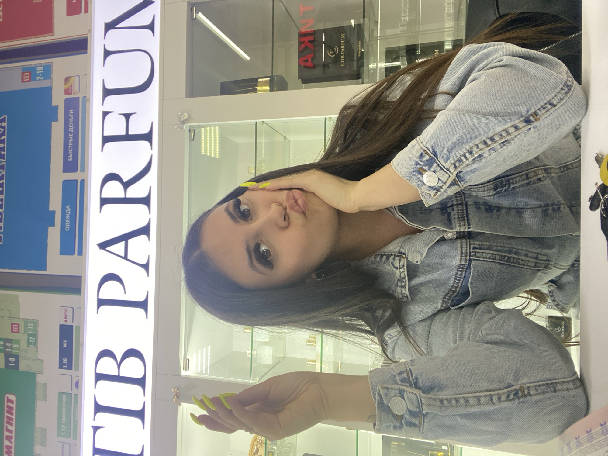 The image is a tilted selfie of a woman with long dark hair, wearing a light blue denim jacket and yellow nail polish. She is making a pouting face and holding her hand near her face. The background appears to be inside a shopping center or mall, with glass display cases filled with perfumes. Behind her, there is a sign that reads "TB PARFUM" in large, illuminated letters. To the left of the sign is a directory with Russian text, including words like "MAGNIT", "ODEZHDA", and "BYSTRIE DENGI". A directory section has "2-10" written on it. Inside a display case "ETIB PARFUM" in black box is visible along with the red "AXINT" logo. The image was likely taken in the city of Chaikovsky, Russia, as the sign and directory appear to be in Russian.