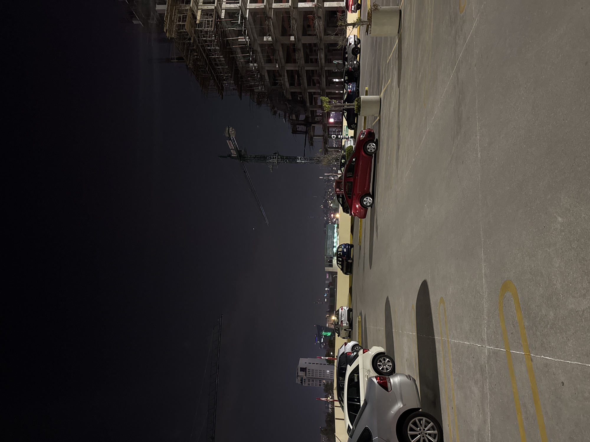 The image shows a nighttime scene in what appears to be a parking structure, possibly in Heroica Puebla de Zaragoza, Mexico, given the city lights in the background. Several cars are parked along the right side of the frame, including a red hatchback, a white SUV, and other dark-colored vehicles. In the distance, a construction site is visible with a crane, and a building under construction is illuminated. The parking structure has yellow parking space lines on the pavement. The sky is dark, suggesting it is either late evening or nighttime. The image is slightly rotated counterclockwise. The location in Heroica Puebla de Zaragoza, Mexico, has buildings under construction and city lights that seem consistent with that area.