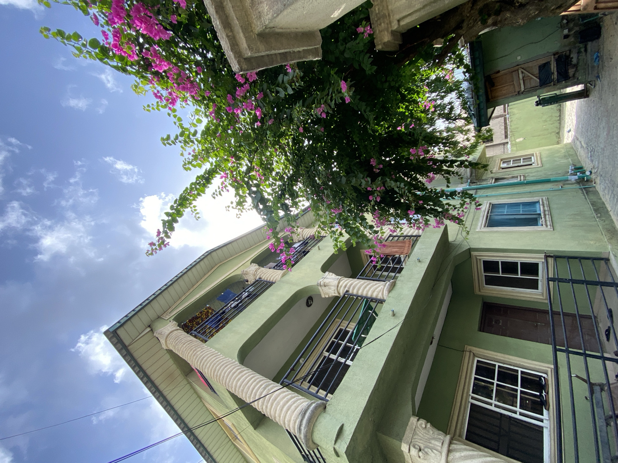 The image depicts a low-angle view of a green, multi-story building with ornate, spiraled column details on the balcony arches. The building appears to be located in an urban area, with a narrow alleyway visible on the right side. A vibrant bougainvillea bush with pink flowers partially obscures the view of the sky, which is blue with scattered clouds. The building has multiple windows and a few balconies. A metal railing is visible on the right side. The location appears to be Ado, Nigeria, based on the provided information. There are no people visible in the scene.