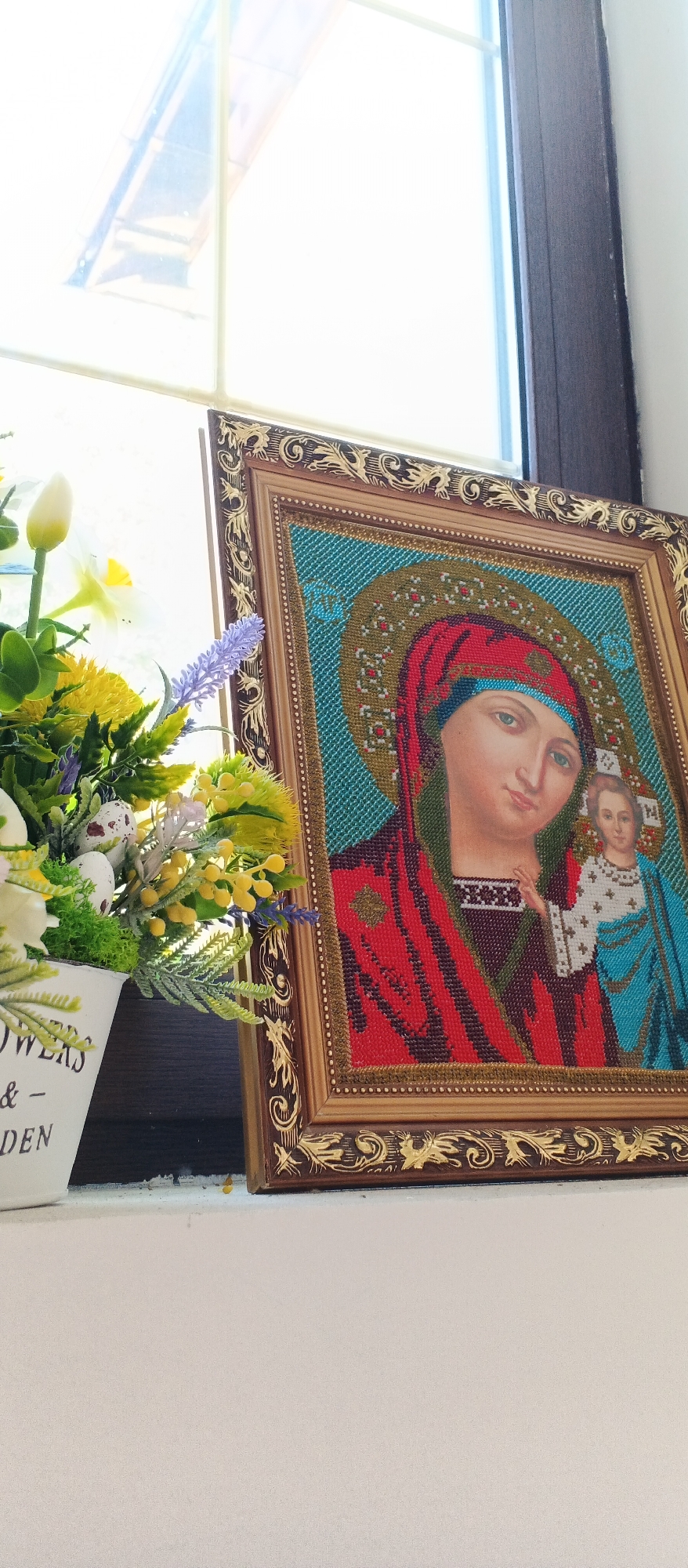 The image shows a close-up, eye-level shot of an icon depicting the Virgin Mary and child Jesus, placed on a windowsill. To the left of the icon is a white planter filled with artificial flowers, predominantly yellow and white, with some green foliage and small decorative eggs. The icon is framed in ornate gold and brown. Behind the objects, a window is visible, with sunlight streaming through, creating a bright, blown-out background. The scene appears to be indoors, on a windowsill of a building in the city of Smila, Ukraine. The specific location within Smila is not identifiable from the image alone.