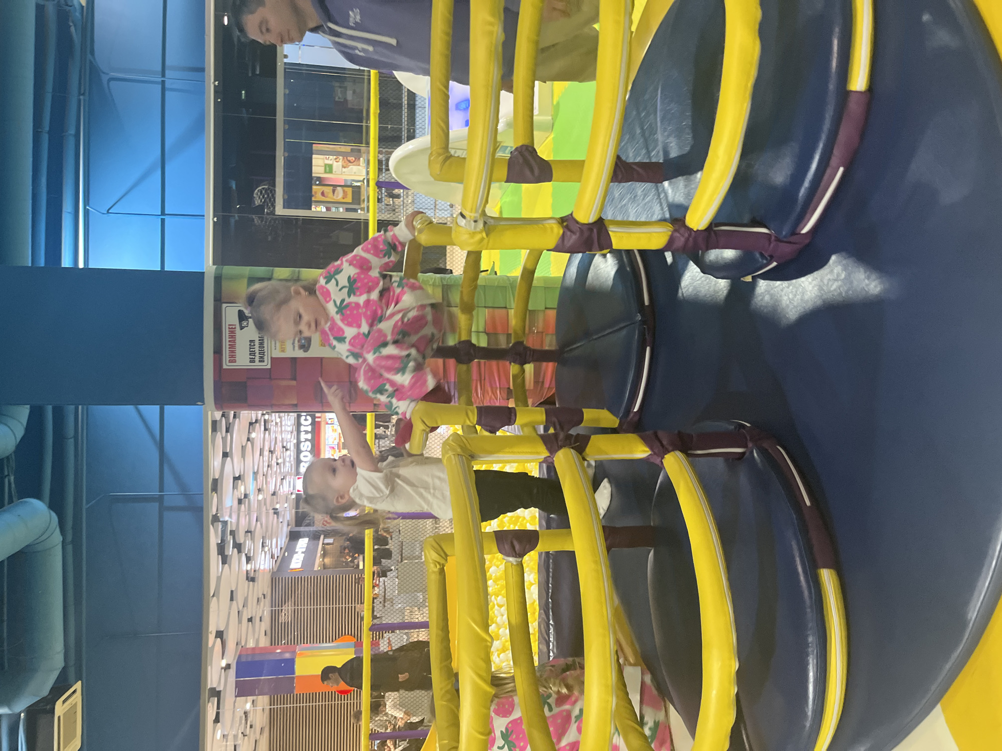 Two young girls are climbing on yellow and blue padded play equipment in an indoor playground, likely at the "MoreMall" shopping center in Sochi, Russia. One girl wears a strawberry-patterned top, and the other wears a white t-shirt and dark pants. An adult is partially visible in the background. The playground includes a ball pit. The background shows a shopping center with stores and white circular decorations on the ceiling. A security camera warning sign is visible on a wall.