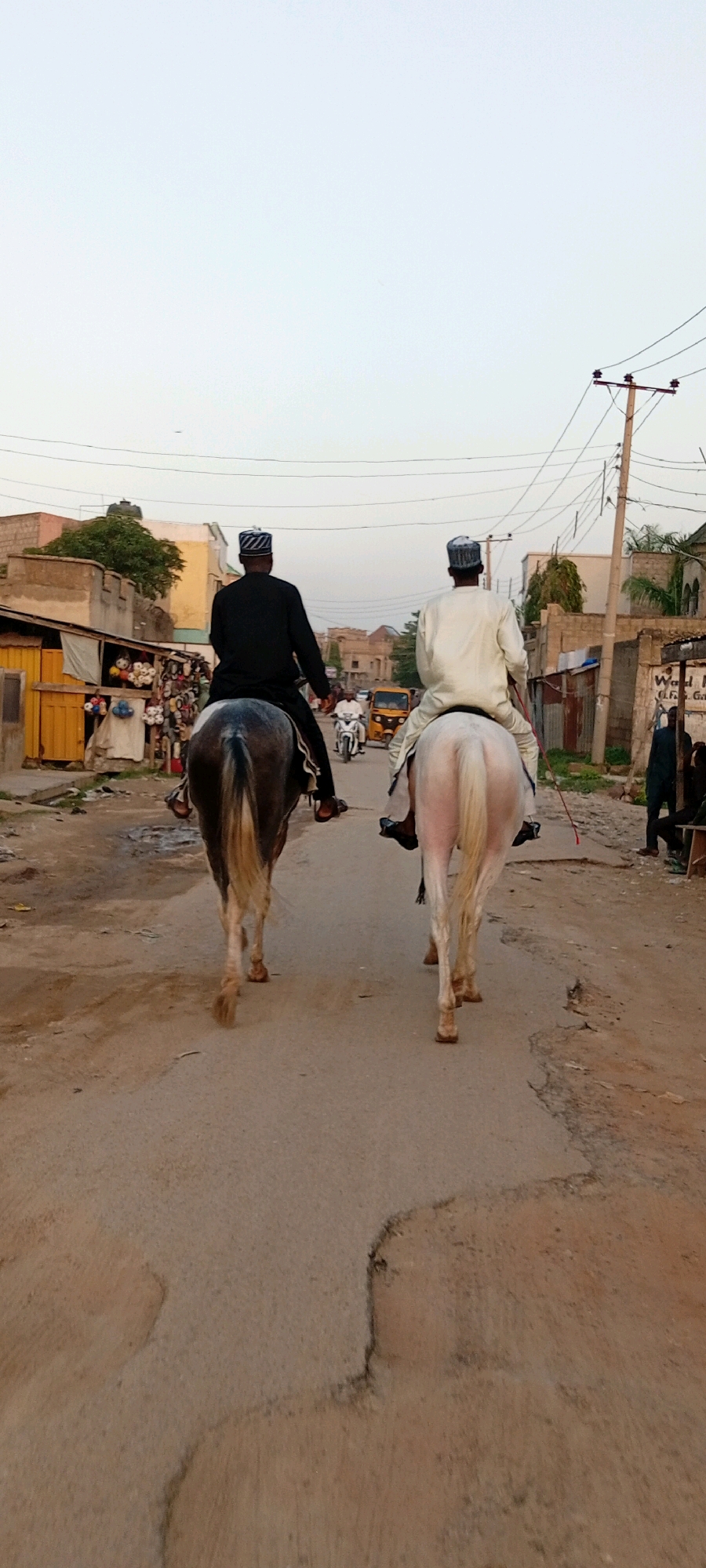 In Kano, Nigeria, two individuals on horseback ride down a dusty, narrow street. The person on the left rides a dark gray and white horse, wearing a black long-sleeved garment and a patterned cap. The person on the right rides a white horse, dressed in a white long-sleeved garment and a patterned cap. Both riders face away from the camera. The street is lined with buildings, some appearing to be shops with goods displayed outside, including colorful textiles. A few motorcycles and a yellow tricycle are visible further down the street, suggesting a bustling urban environment. Power lines and poles are present in the background. The sky is a pale, overcast gray.