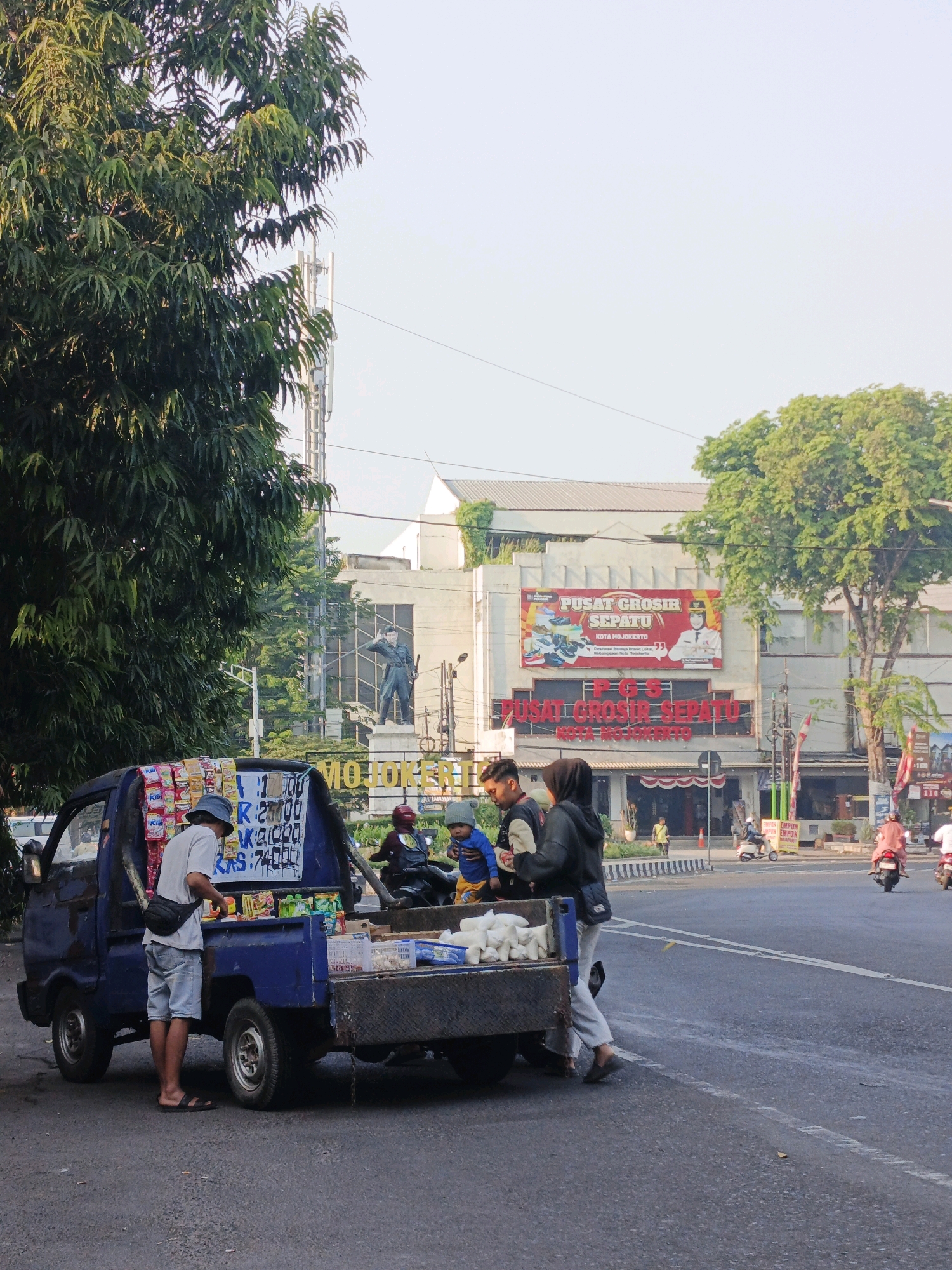 A blue pickup truck, converted into a mobile merchandise stand, is parked on the side of a street in Kranggan, Indonesia. The truck is laden with various goods, including packaged snacks and drinks displayed on its side. A man in a blue hat and shorts stands on the left, interacting with the stand. To the right, two people, one wearing a hijab, are near the back of the truck, possibly making a purchase or observing the goods. The background reveals a building with a large banner reading "PUSAT GROSIR SEPATU" (Shoe Wholesale Center) and a statue of a soldier. Other pedestrians and a few motorcycles are visible on the street, indicating a moderately busy urban environment. The lighting suggests it is daytime, with clear skies.