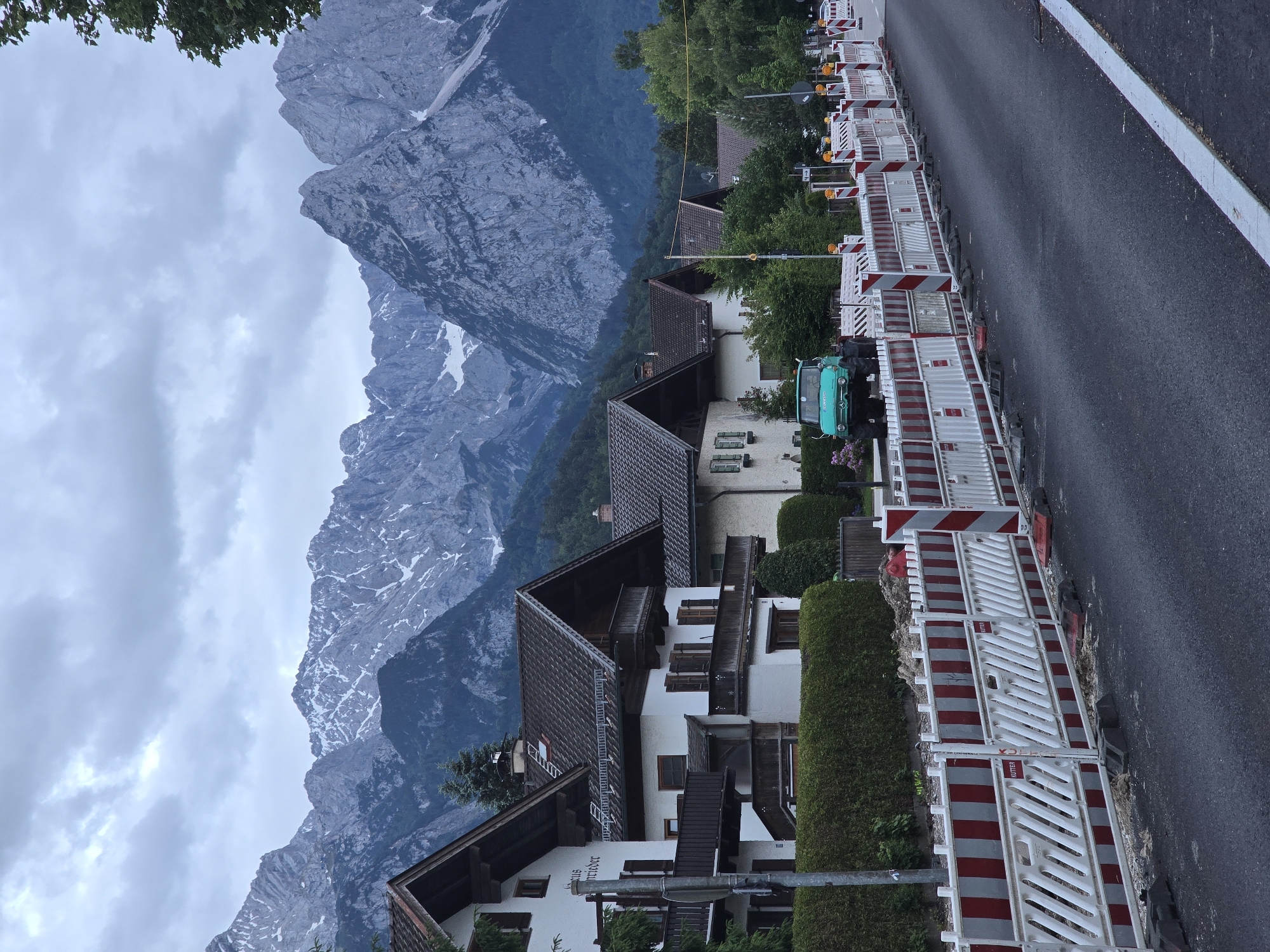 This image depicts a street scene in Garmisch-Partenkirchen, Germany, characterized by residential architecture set against a mountainous backdrop.
In the background, a range of large, jagged mountains dominates the horizon under an overcast sky. Patches of snow are visible on the upper slopes and peaks of the mountains, which transition to darker forested areas at lower elevations.
The middle ground features a row of multi-story traditional German houses, predominantly white or light-colored, with steep, dark-tiled gable roofs, wooden balconies, and dark shutters. The first house on the left has "Haus Schreieder" written on its facade. A well-maintained green hedge runs in front of these houses. A distinctive green Mercedes-Benz Unimog utility vehicle is parked partially obscured by trees and a hedge on the left side of the street. Utility poles and overhead lines are also present.
The foreground is occupied by a paved road, which appears to be under construction or recently repaved, given its smooth, dark surface and the presence of numerous temporary red and white construction barriers. These barriers line the right side of the road, extending into the distance, with some featuring orange flashing lights (though not active). Visible on the barriers is the text "KUTTER," indicating the construction company. A white solid line marks the right edge of the road, which curves slightly to the right in the distance. No people are visible in the scene.