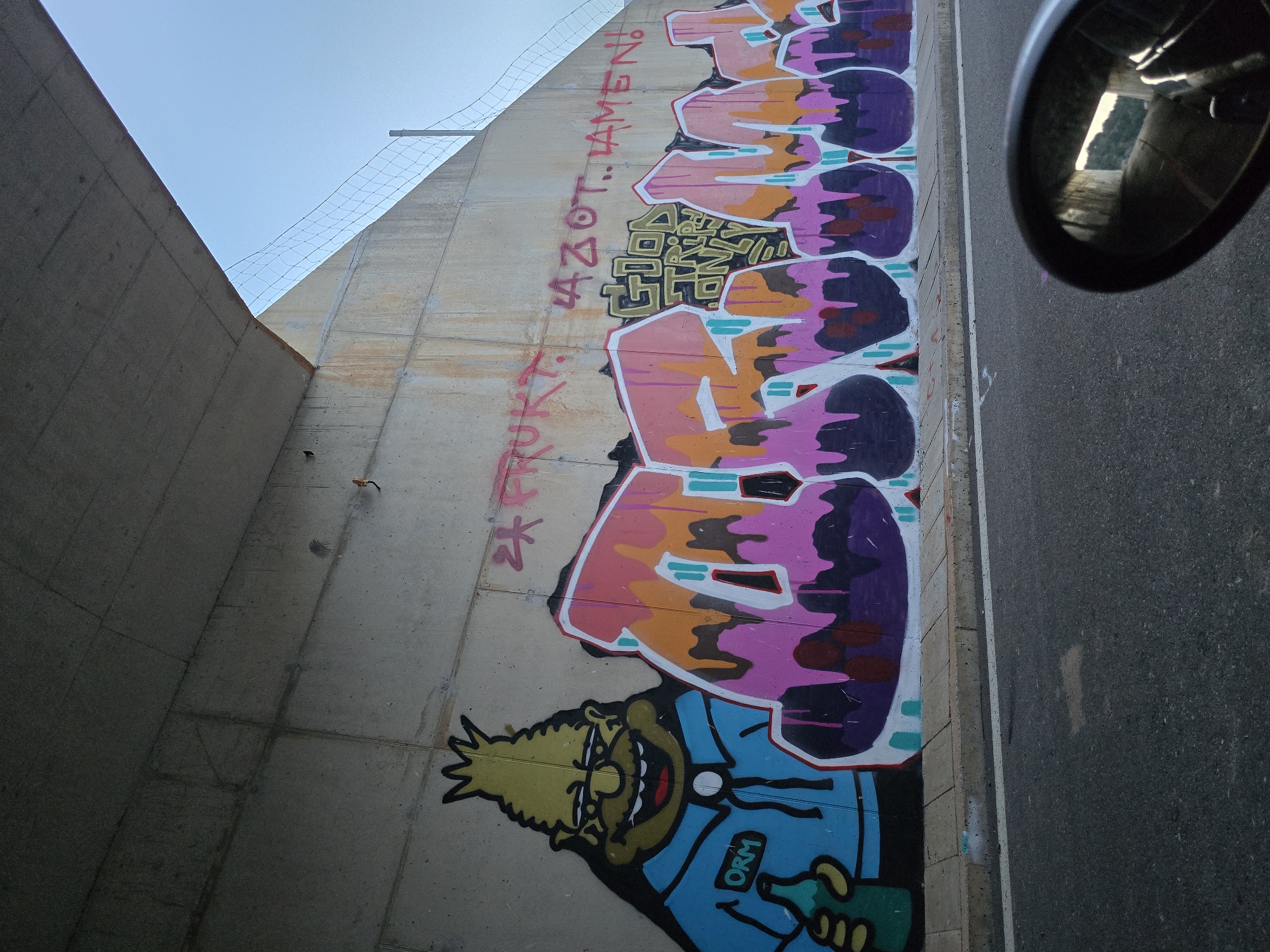 The image captures a section of a concrete underpass in Klopot, Montenegro, adorned with graffiti. On the left, a cartoonish character with spiky yellow hair, glasses, and a blue shirt emblazoned with "ORM" holds a green bottle. To its right, large, vibrant graffiti letters in pink, orange, purple, and white spell out "DREAM". Within the "E" of "DREAM" is another, smaller piece of graffiti in yellow that reads "GOOD TRIPS ONLY". Above the large graffiti, in smaller red lettering, is the text "24 PRUKT AZOT. LAMEN!". The underpass wall is concrete, showing some weathering and stains. A road runs along the bottom of the frame, with a curb and a white line marking the edge. In the lower right corner, a motorcycle's rearview mirror reflects the interior of the underpass and a distant, bright opening. The sky above is a pale blue.
