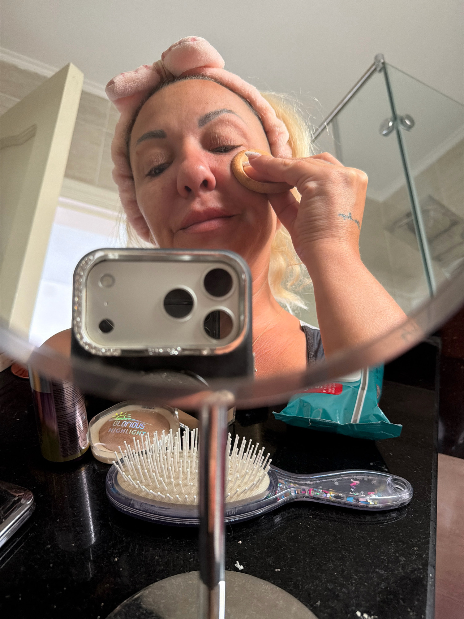 A woman with blonde hair, wearing a pink headband, is applying makeup in a bathroom. She is holding a makeup sponge to her right cheek, near her eye. Her eyes are closed. A mirror reflects her face and the surrounding environment. On the counter in front of her, there are several makeup items, including a compact with "GLORIOUS HIGHLIGHT" written on it and a hairbrush. A partially visible smartphone case with a camera array is also reflected in the mirror. In the background, a glass shower enclosure is visible. The scene takes place in a modern bathroom, likely in a hotel in Sharm El Sheikh, Egypt, judging by the tiles on the wall and the overall aesthetic.