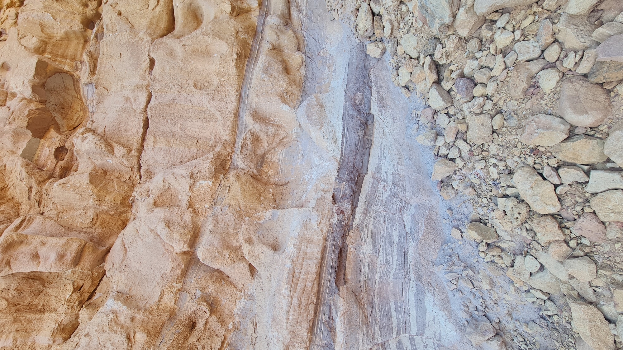 The image depicts a close-up view of a rock formation in Hevel Eilot Regional Council, Israel. The formation is composed of three distinct sections. The leftmost section is a rough, textured rock face with a tan color. The central section is smoother and lighter in color, with visible vertical striations. The rightmost section consists of loose rocks and pebbles, also in a tan color. The composition is a ground-level shot, focusing on the texture and variation in the rock. There are no people visible.