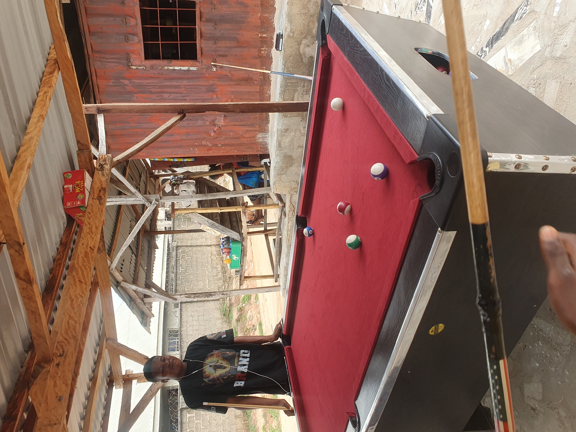 In Lokoja, Nigeria, a young Black male is standing next to a red-felted pool table, holding a pool cue. He wears a black t-shirt with a graphic design and white earbuds. The pool table has several balls on it, including a striped blue and white ball, a solid green ball, a solid red ball, and a solid white ball. The scene appears to be outdoors, under a rustic, corrugated metal roof supported by wooden beams. In the background, there are brick walls, a rusty metal structure with a window, and various items, including bottles and a green crate on a wooden table. The ground is a mix of concrete and sand. A pool cue with an American flag motif is visible in the foreground, obscuring part of the pool table.