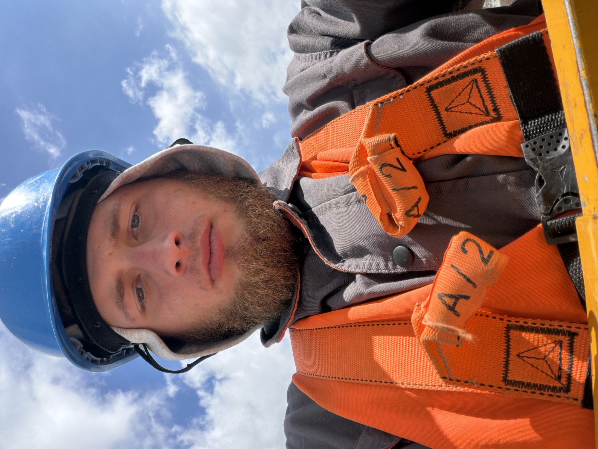 A close-up, low-angle selfie captures a young man wearing a blue hard hat and an orange safety vest over a gray work shirt. The man has a light brown beard and is looking directly at the camera with a neutral expression. The safety vest has straps with the marking "A/2" and a triangular logo. The background consists of a bright blue sky with scattered white clouds, suggesting an outdoor setting. The image was taken in Stróża, Poland.
