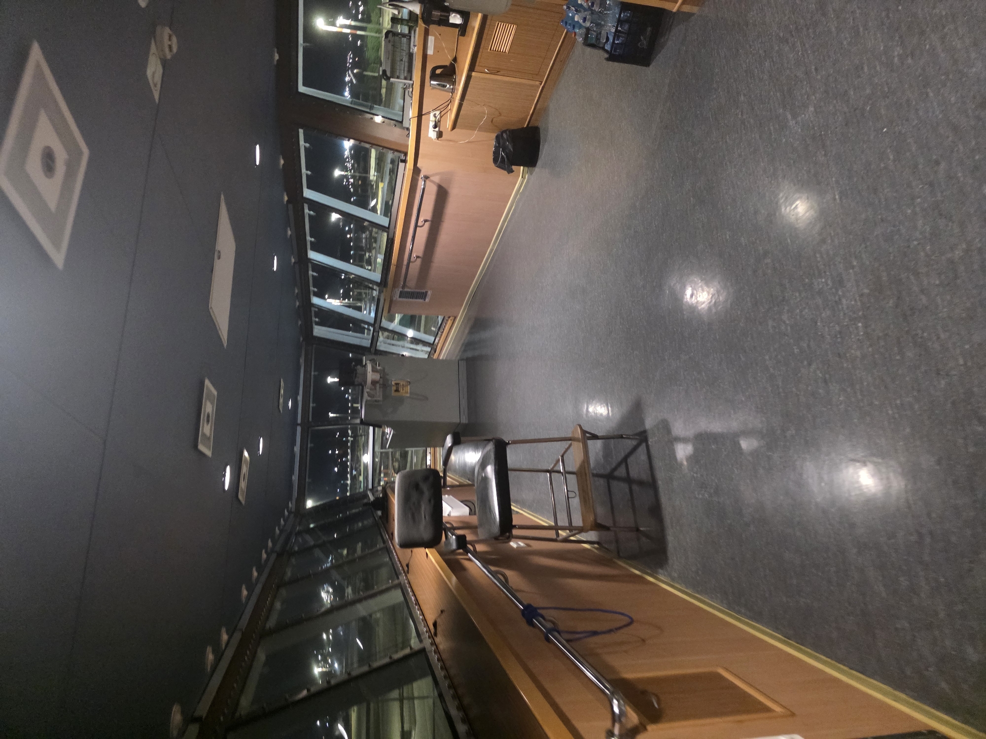 The image depicts the interior of a vessel's bridge or control room at night. The room is spacious with a dark gray ceiling containing recessed lighting and square decorative panels. Large windows line the perimeter, offering a view of a brightly lit cityscape or port, possibly Brindisi, Italy, with numerous streetlights and building lights visible.
In the foreground, a dark gray, textured floor reflects some of the overhead lighting. To the left, a wooden paneling lines the wall, with a chrome handrail attached. A blue rope is looped through the handrail. A black leather captain's chair with a metal frame and wooden footrest stands near the wall.
Further into the room, a lighter-colored wooden cabinet unit is visible along the right wall. On top of this unit, there is a black trash can, an electric kettle, and what appears to be a coffee maker. Next to the cabinet, a crate filled with bottled water is placed on the floor. The overall impression is of a functional, dimly lit operational space within a vessel.