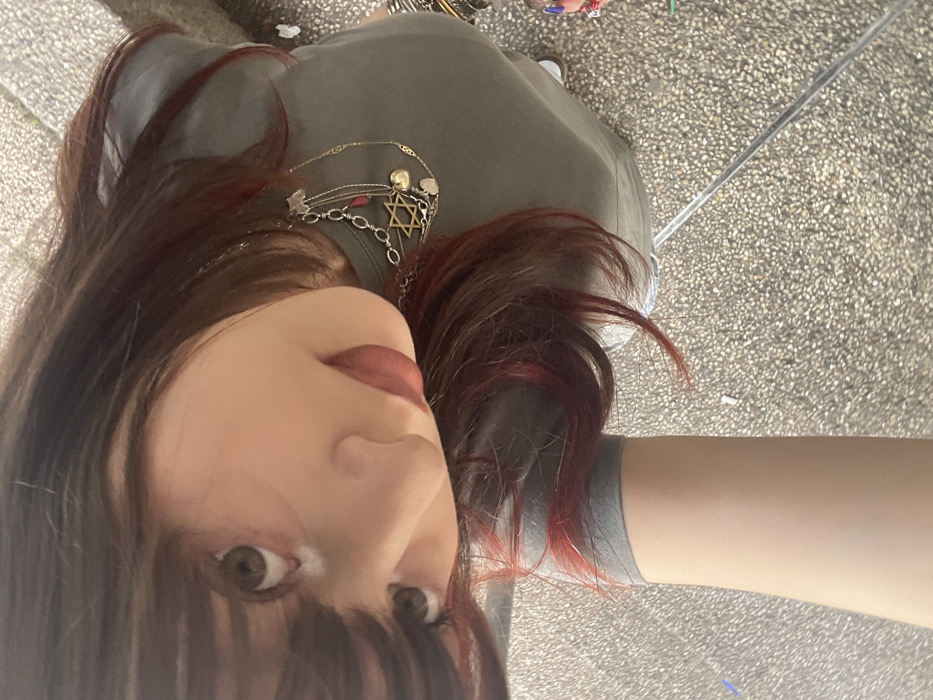 A person, identified as female, is depicted in an upward-looking close-up, gazing directly at the camera. She possesses dark brown hair with reddish highlights and bangs, and wears an olive-green short-sleeved t-shirt. Dark eyes and lips colored with a dark pink/mauve lipstick are prominent. Multiple layered necklaces adorn her neck: one features a gold-toned Star of David pendant on a silver-toned chain, another is a thin gold-toned chain with small charms, and a third silver-toned chain displays assorted smaller charms, including a red bead. A portion of her left forearm extends into the lower frame. The background consists of a textured, light grey and white speckled paved surface, characteristic of an outdoor pedestrian area in an urban or suburban environment. A faint diagonal reflection, possibly from a glass surface, crosses the lower right. Outdoor daytime lighting illuminates the scene. This location is identified as being within the city of Raanana, Israel.