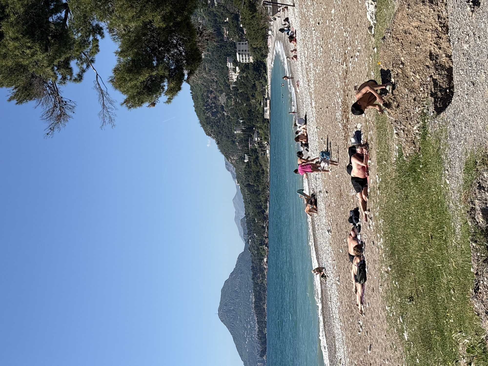 The image shows a beach in Bar, Montenegro. The beach is made of small pebbles and is populated with people sunbathing, swimming, and walking. The sea is turquoise and clear, with small waves breaking on the shore. A mountainous landscape covered in lush greenery forms the backdrop. A few buildings dot the mountainside. The sky is clear and blue, with a few tree branches visible in the upper left corner. A person is digging a hole in the sand near the edge of a patch of grass.