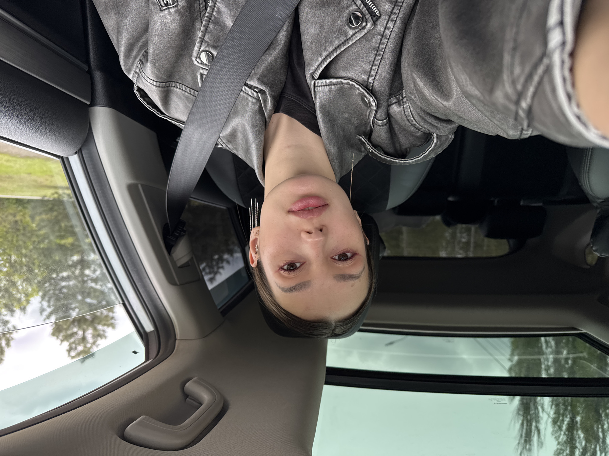 A selfie taken inside a car. A young woman wearing a gray leather jacket and a black T-shirt is looking at the camera. She is wearing a seatbelt. The interior of the car is visible, including the roof and window. The location is unknown.