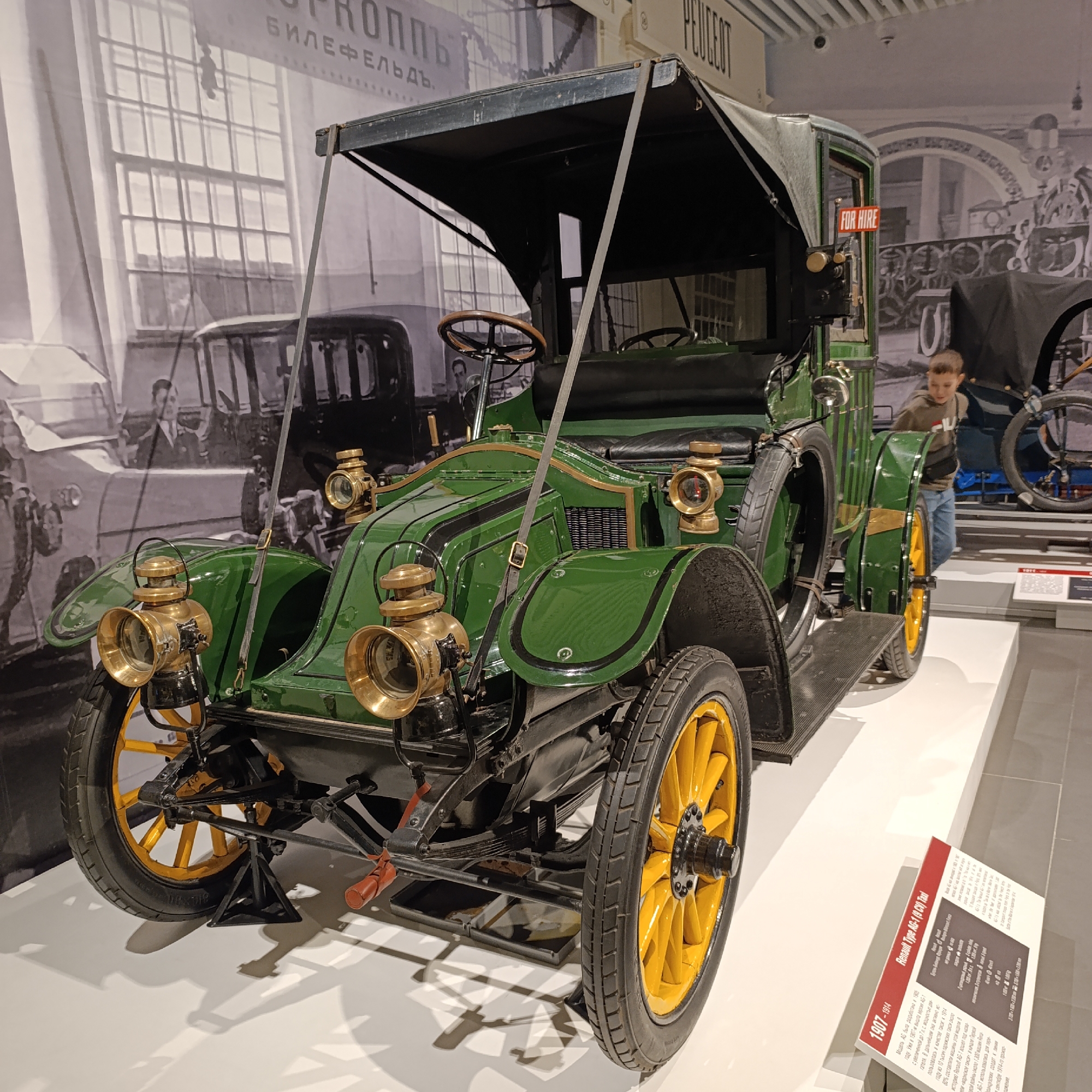 The image shows a vintage green Renault Type AG-1 (9 CV) Taxi from 1907-1914 displayed in a museum setting, specifically in Verkhnyaya Pyshma, Russia. The car has yellow wheels, brass lamps, and a black convertible top. A sign with information about the Renault Type AG-1 (9 CV) Taxi is visible next to the car. A young boy is standing near the car. The backdrop features a black and white photo of early 20th-century vehicles and figures, as well as Russian text "БИЛЕФЕЛЬДъ". A "FOR HIRE" sign is on the vintage car.