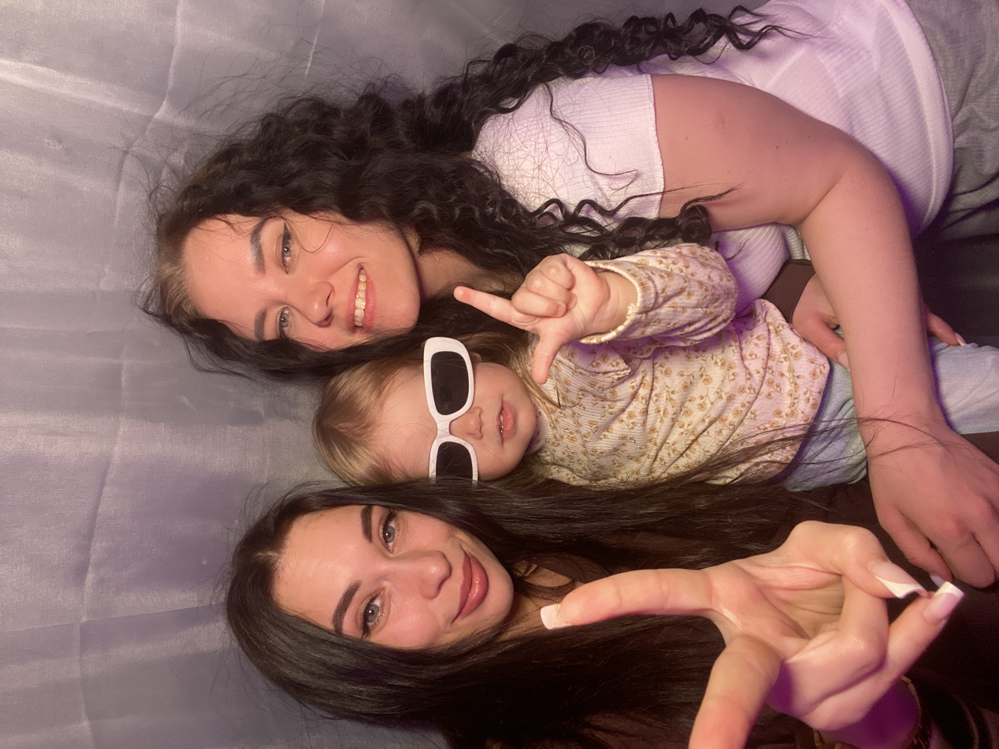 Three individuals are lying down on a gray textured surface, potentially a bed or couch. At the top of the image is a woman with long, dark, curly hair wearing a white ribbed top. A young child wearing white-framed sunglasses and a floral-patterned shirt lies beneath her. The child is making a hand gesture. At the bottom of the image, another woman with long, dark, straight hair is visible. Her hand is raised with her index finger extended. The location is identified as the city of Chaikovsky, Russia. No specific building or landmark is apparent within the frame.